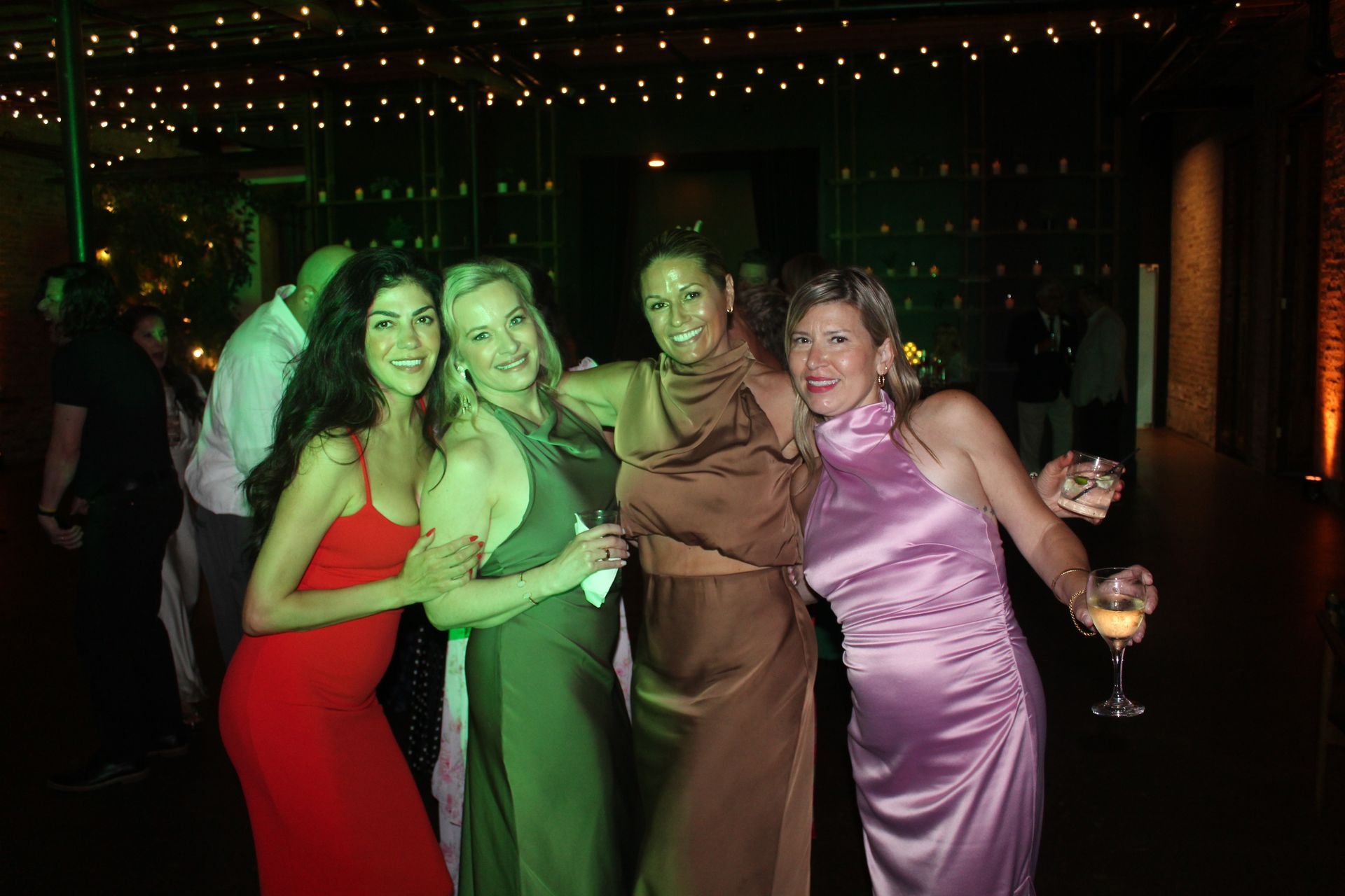Four women in formal gowns pose together at a party with string lights.