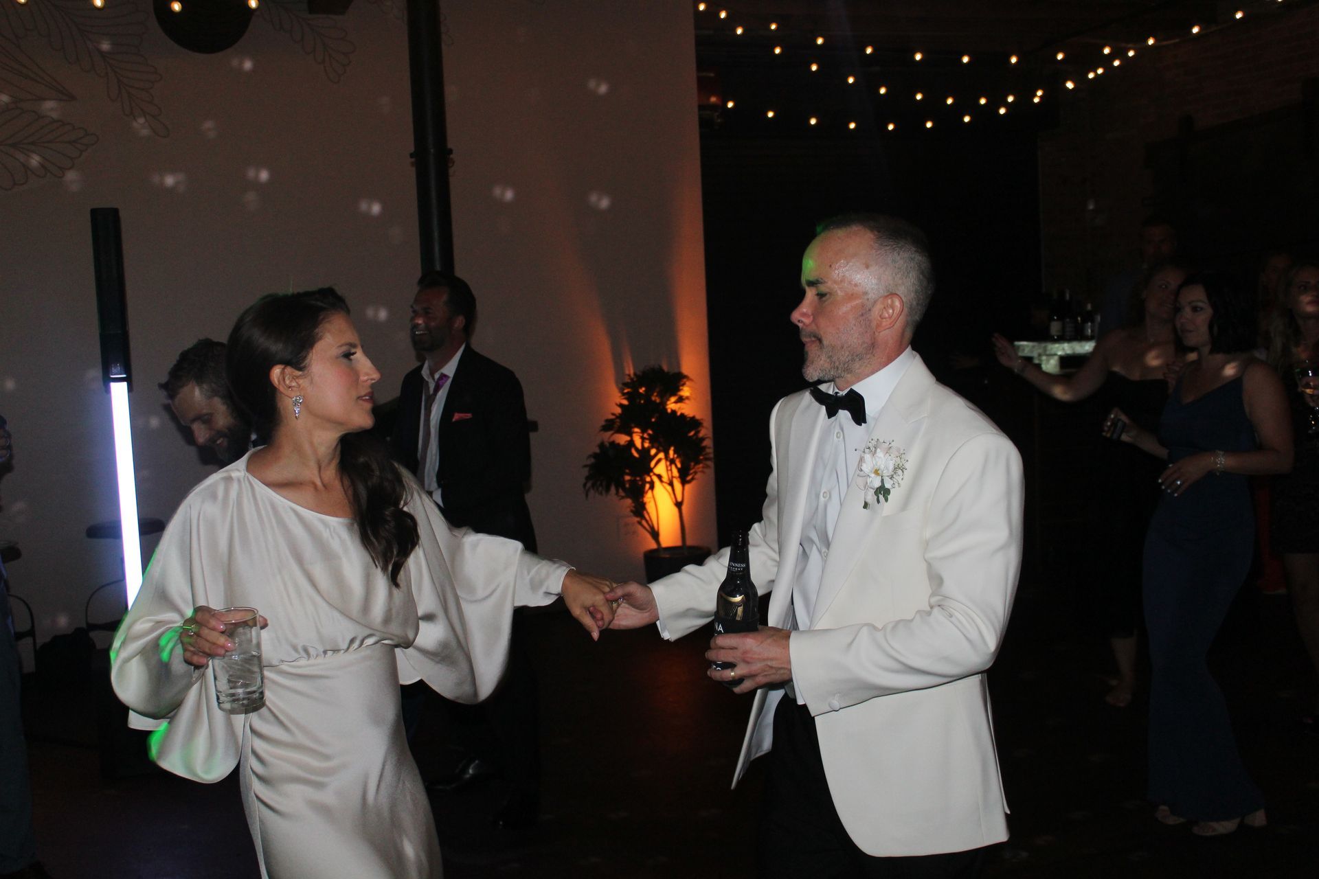 Couple dancing at a reception, the woman in a white dress, the man in a white blazer and black bow tie, lit by disco lights.