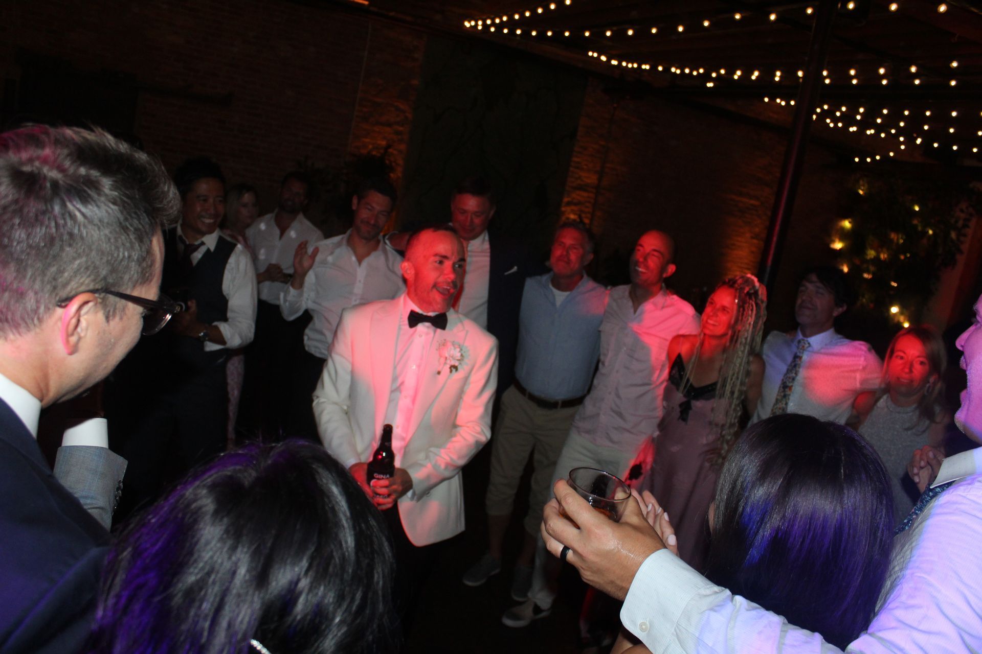 People dancing at an indoor event, under string lights. Some holding drinks, several looking at camera.