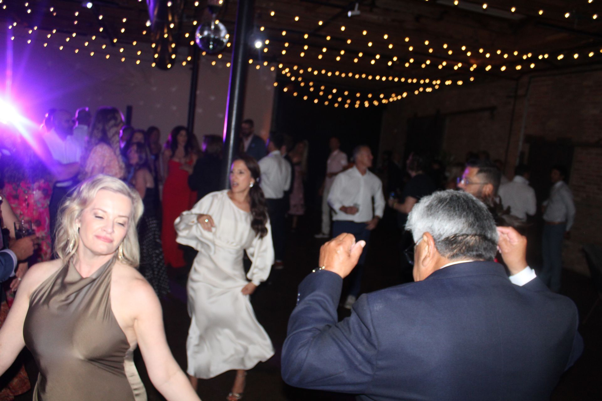 People dancing in a brick-walled room lit by string lights and a disco ball. Some are wearing formal attire.