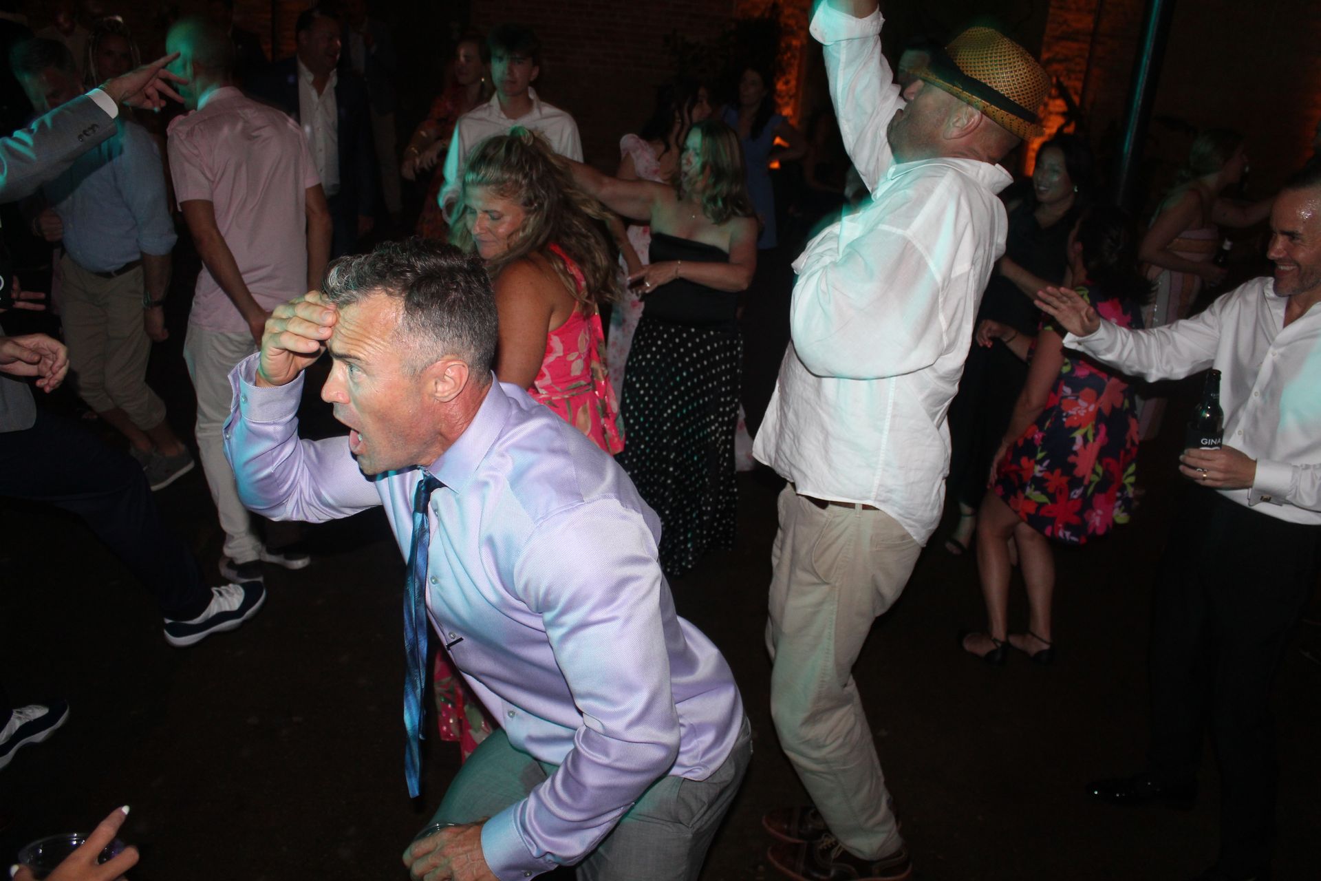 People dancing energetically at a party, illuminated by blue and green lights; one man with a tie bends forward.