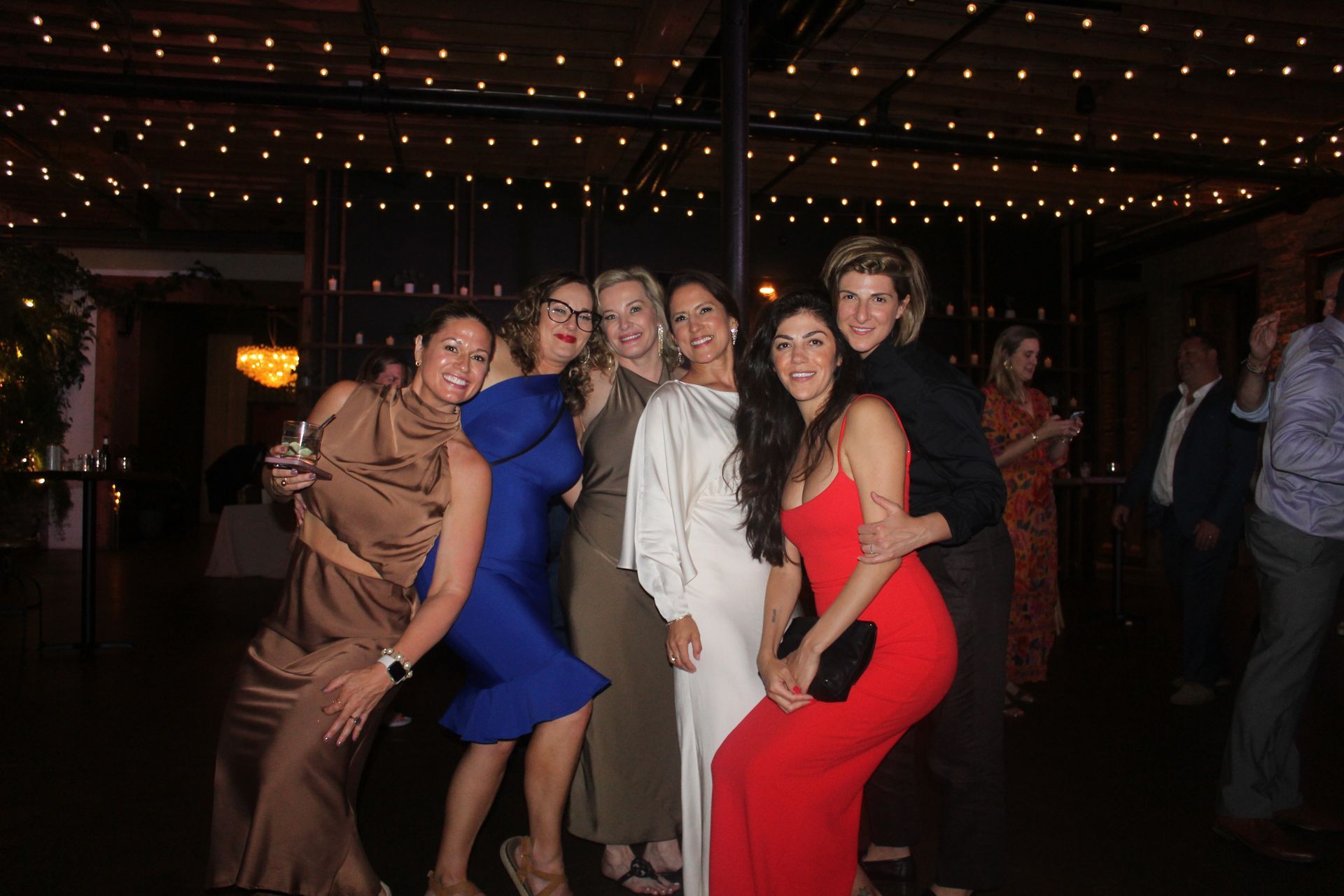 Group of people pose for a photo at an event, smiling, with string lights overhead.