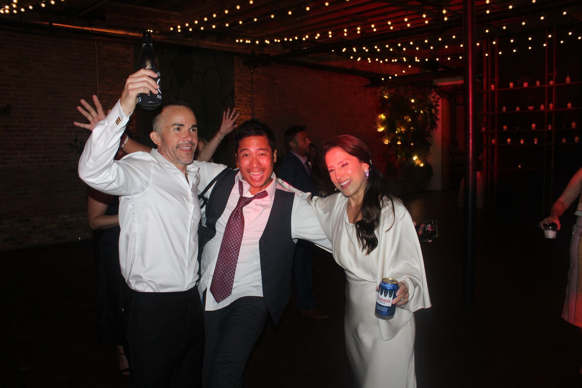 Three people celebrating at a party, holding drinks, under string lights.