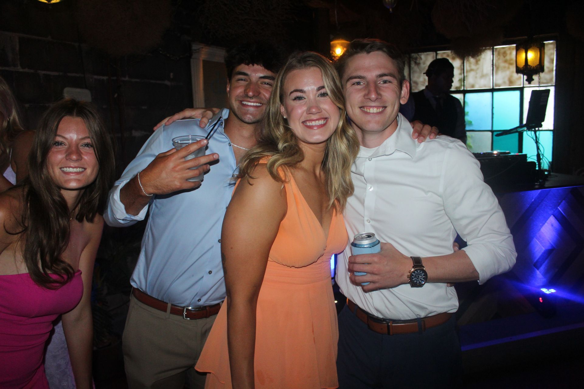 Group of four people smiling and posing in a dimly lit setting, one holding a drink, another a can.