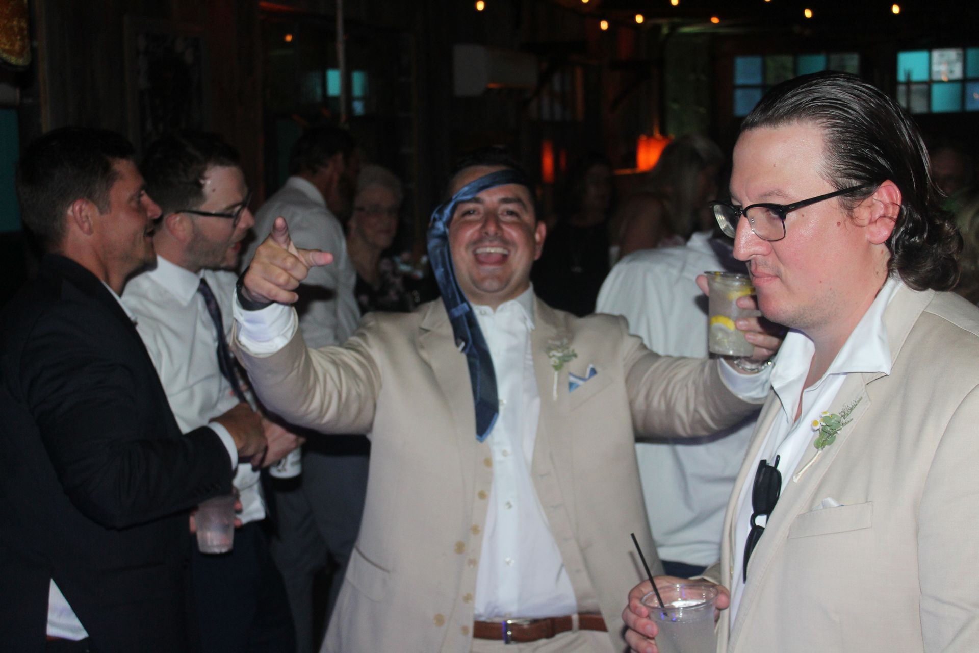 People celebrating in a room. Man in beige suit gestures with tie around neck, smiling. Others near.