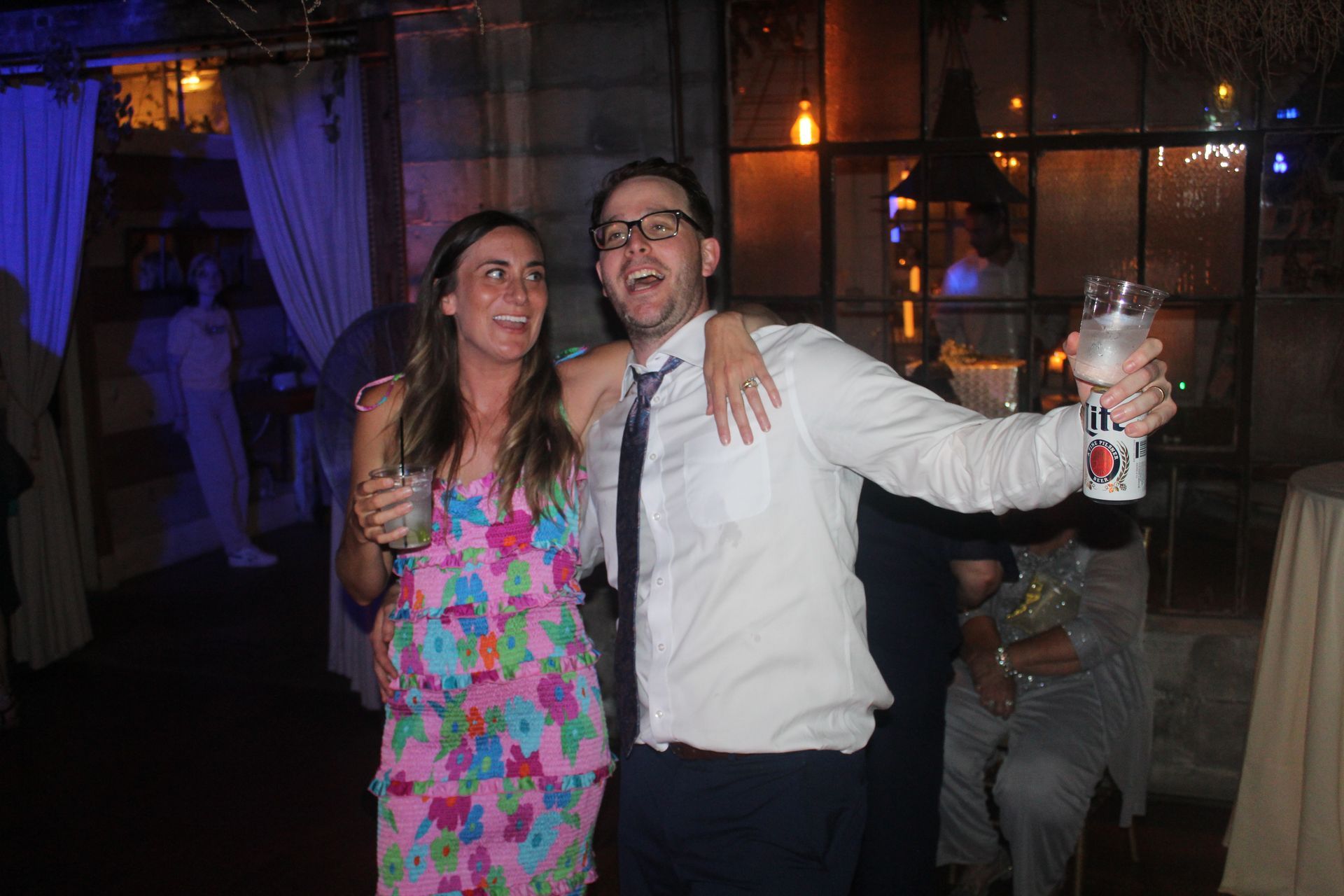 Woman in floral dress and man in a shirt and tie, arm-in-arm, smiling and holding drinks at a dimly lit party.