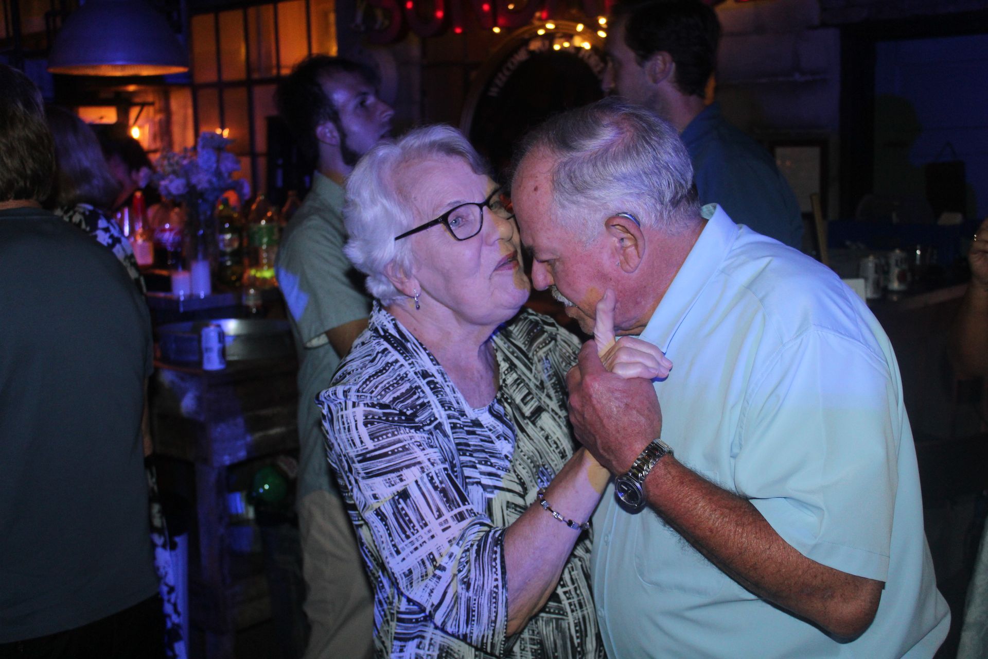 Elderly couple dancing closely at a dimly lit party, man in light blue shirt, woman in patterned top.