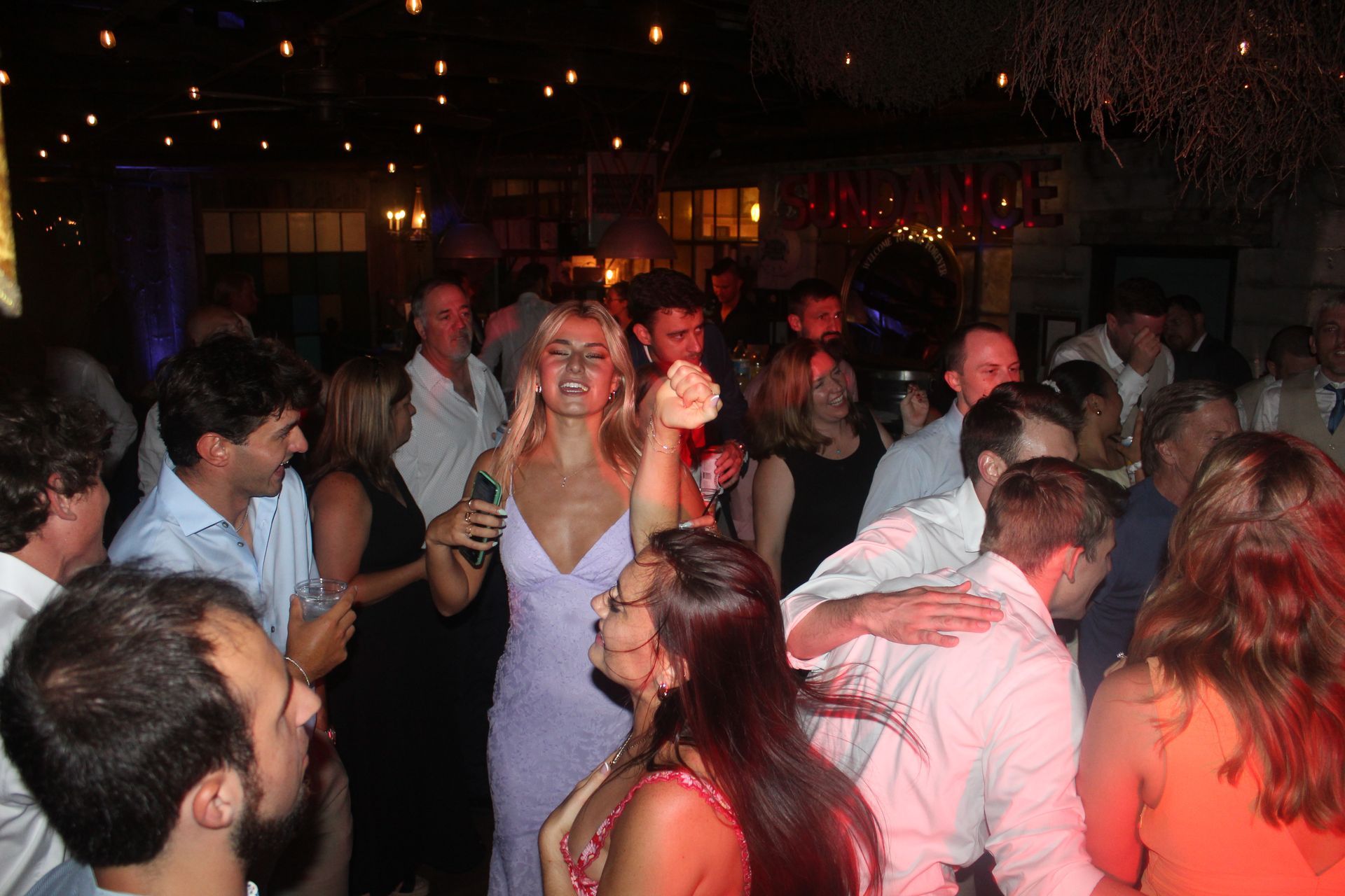 People dancing in a crowded venue, illuminated by overhead lights and red stage lighting.