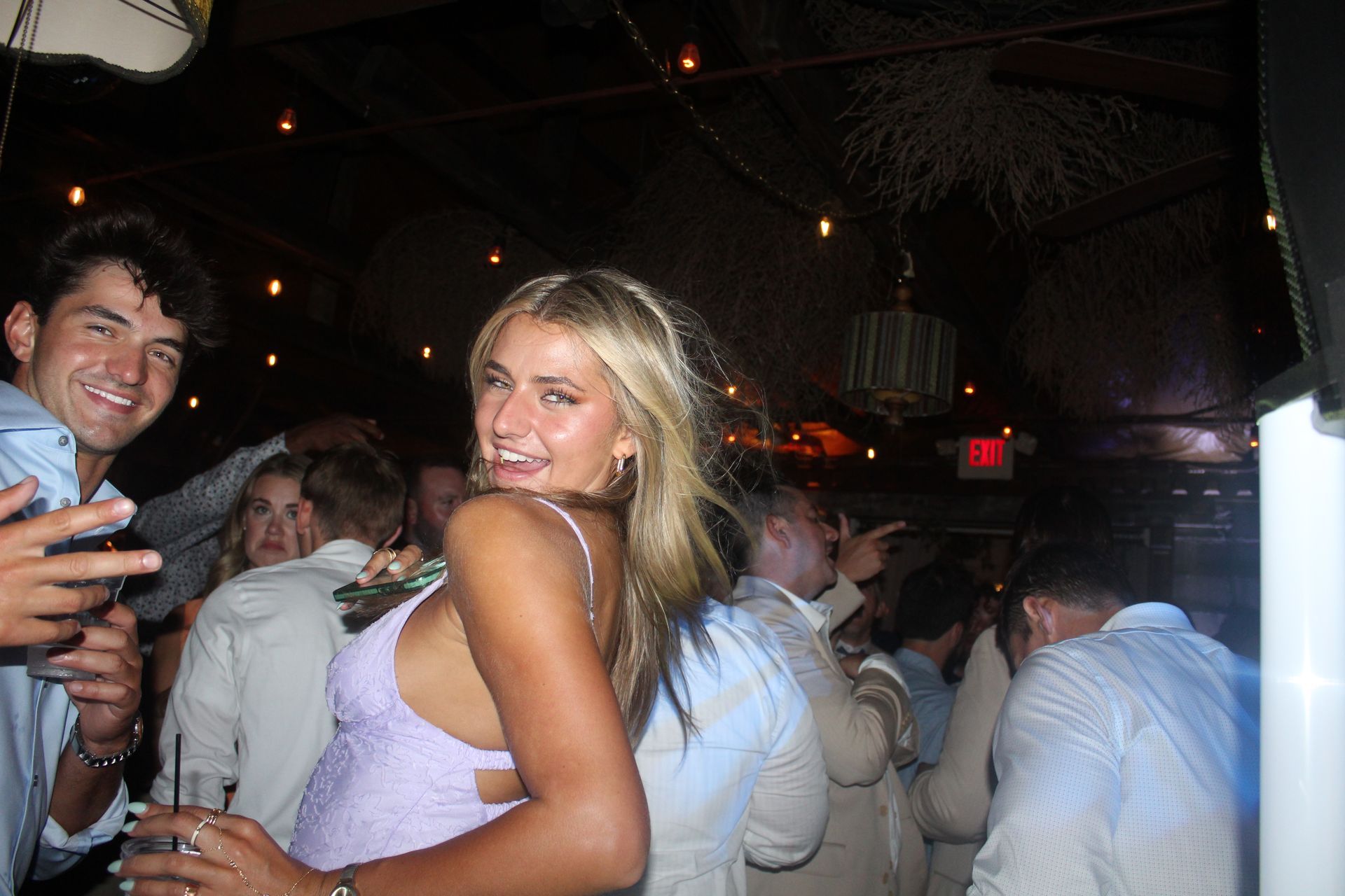 Woman in light purple dress smiles, looks back at camera. Man to her left smiles and points. People in the background.