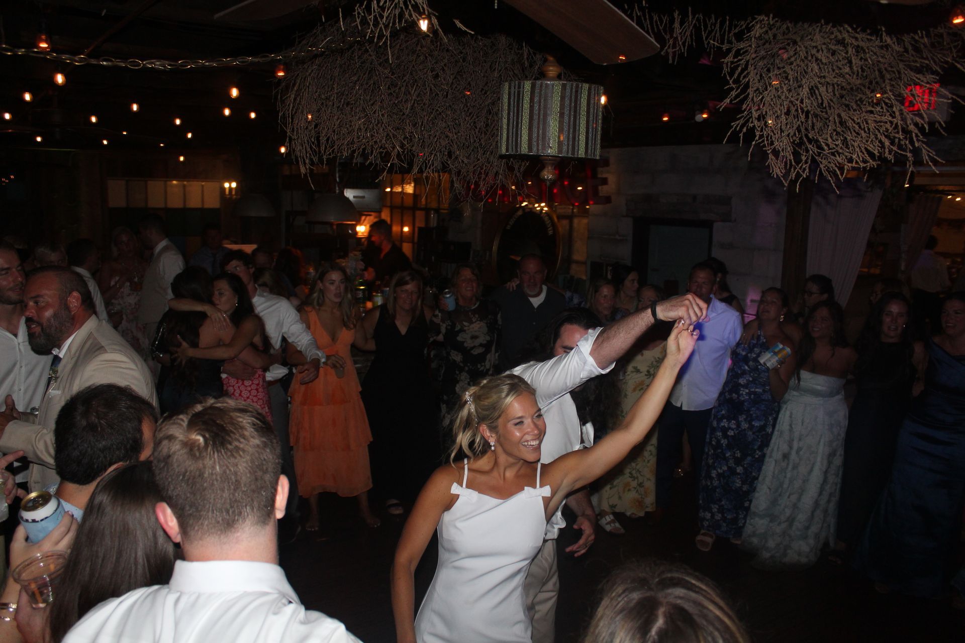 People dancing at an event, inside a venue with string lights and decorations.