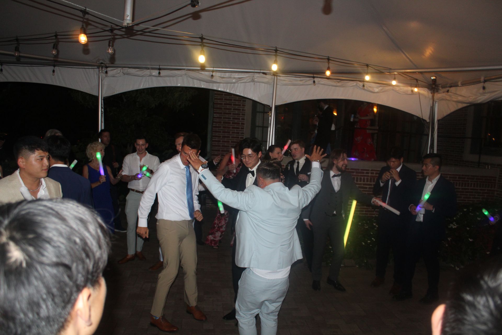 People dancing at an outdoor event under a tent, some holding glow sticks.