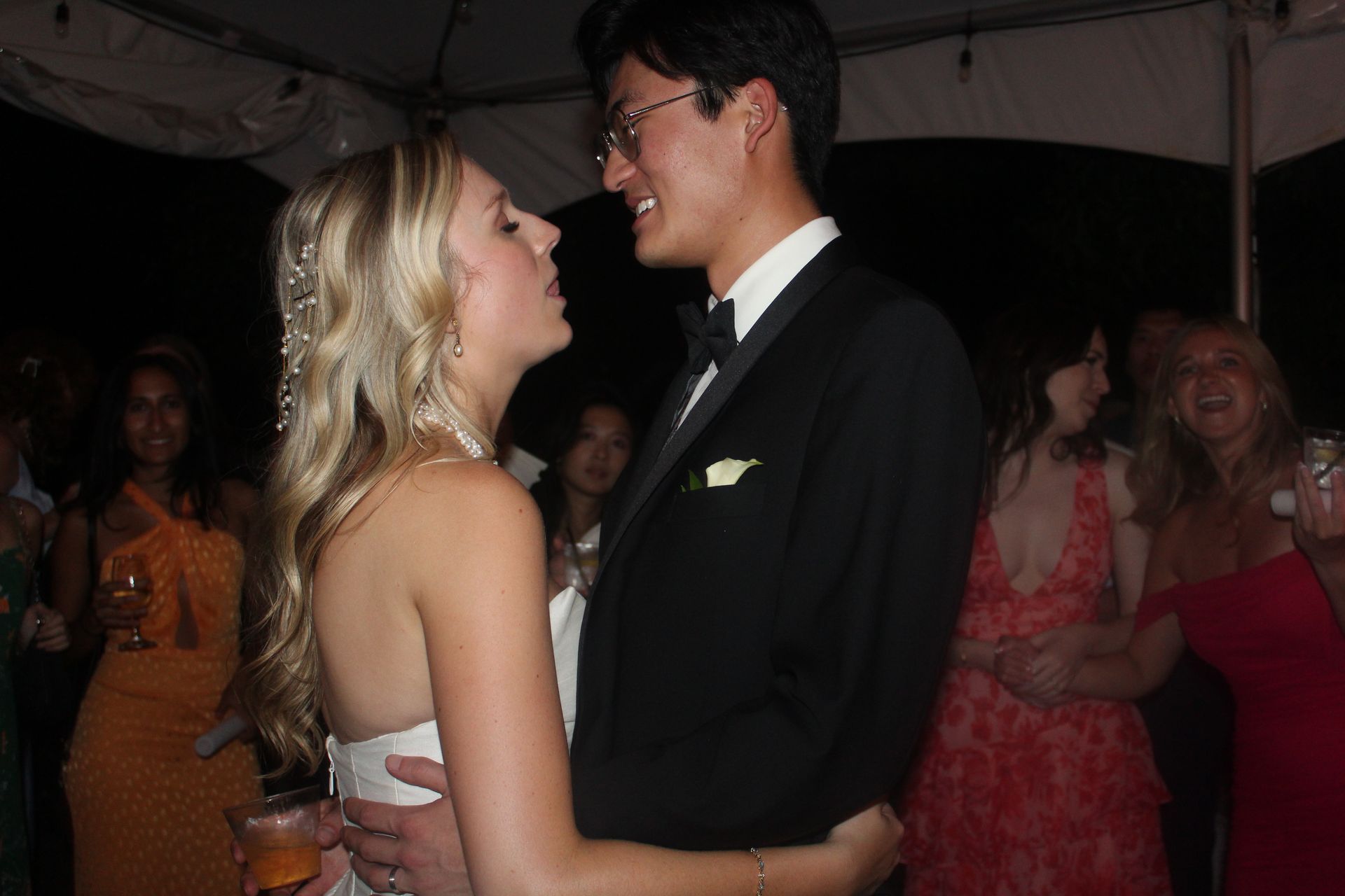 Couple embraces at a celebration, dark formal attire. Women in colorful dresses, string lights in the background.