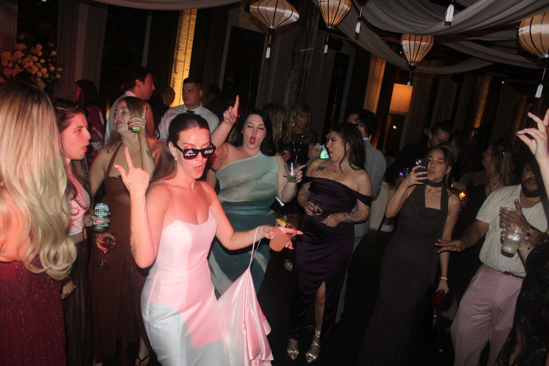 People dancing at a party in a decorated room with drinks, with a woman in a pink dress in the foreground.