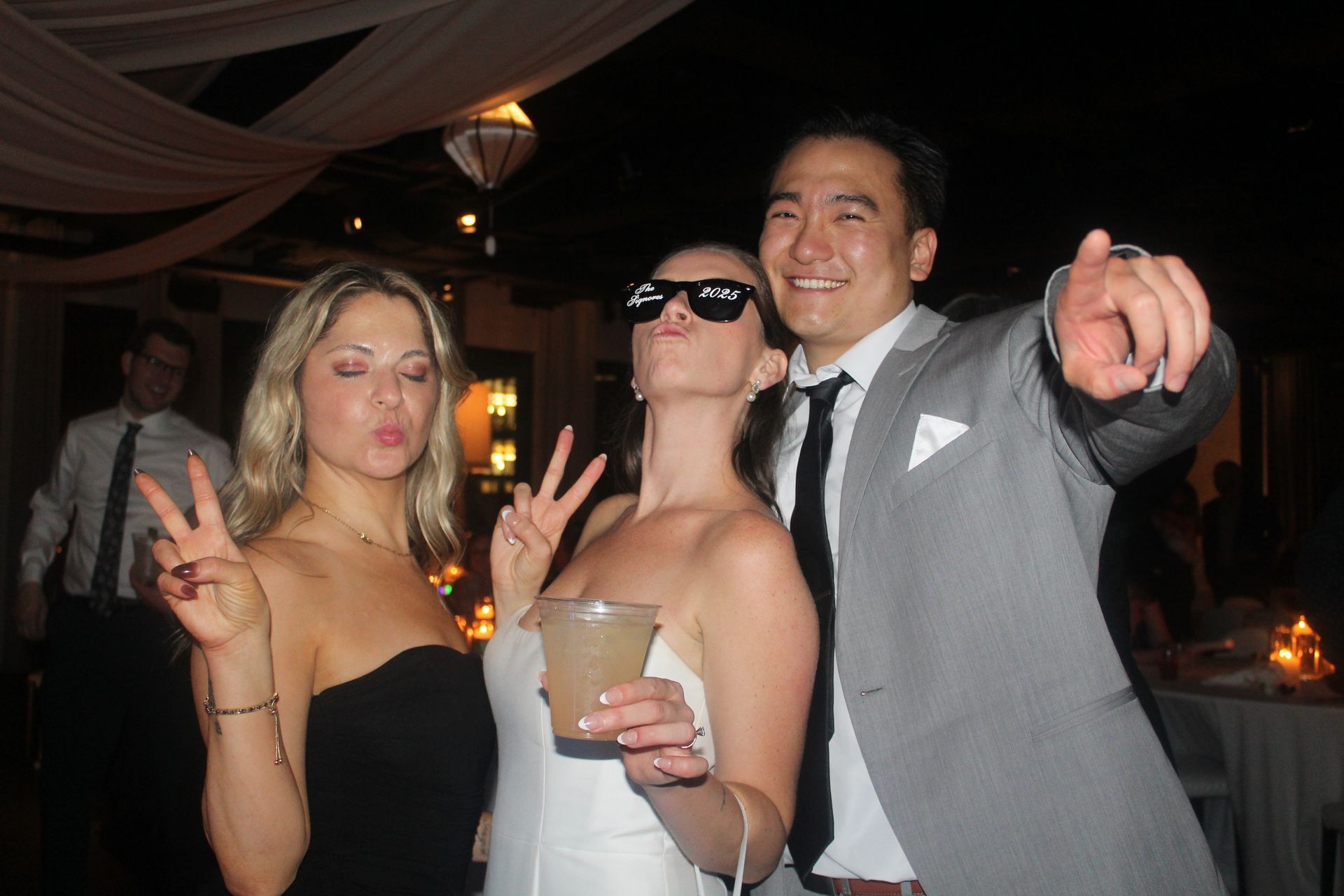 Three people pose for photo at party; two women and one man, making peace signs, man points.