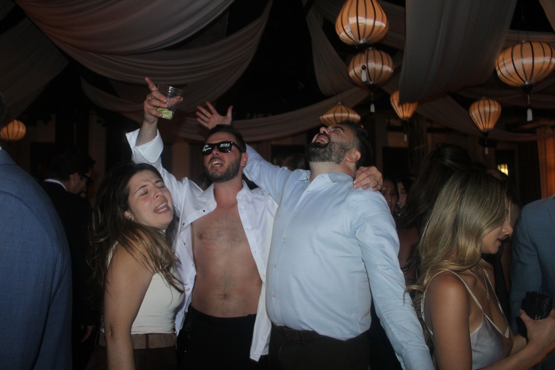 People dancing at a party; man with open shirt raises a drink, another has a cookie on his head. Dim lighting.