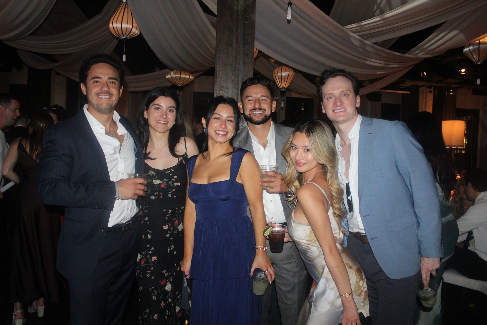 Group of six people smiling at a party with white draping; dark clothing, drinks held, and lights.