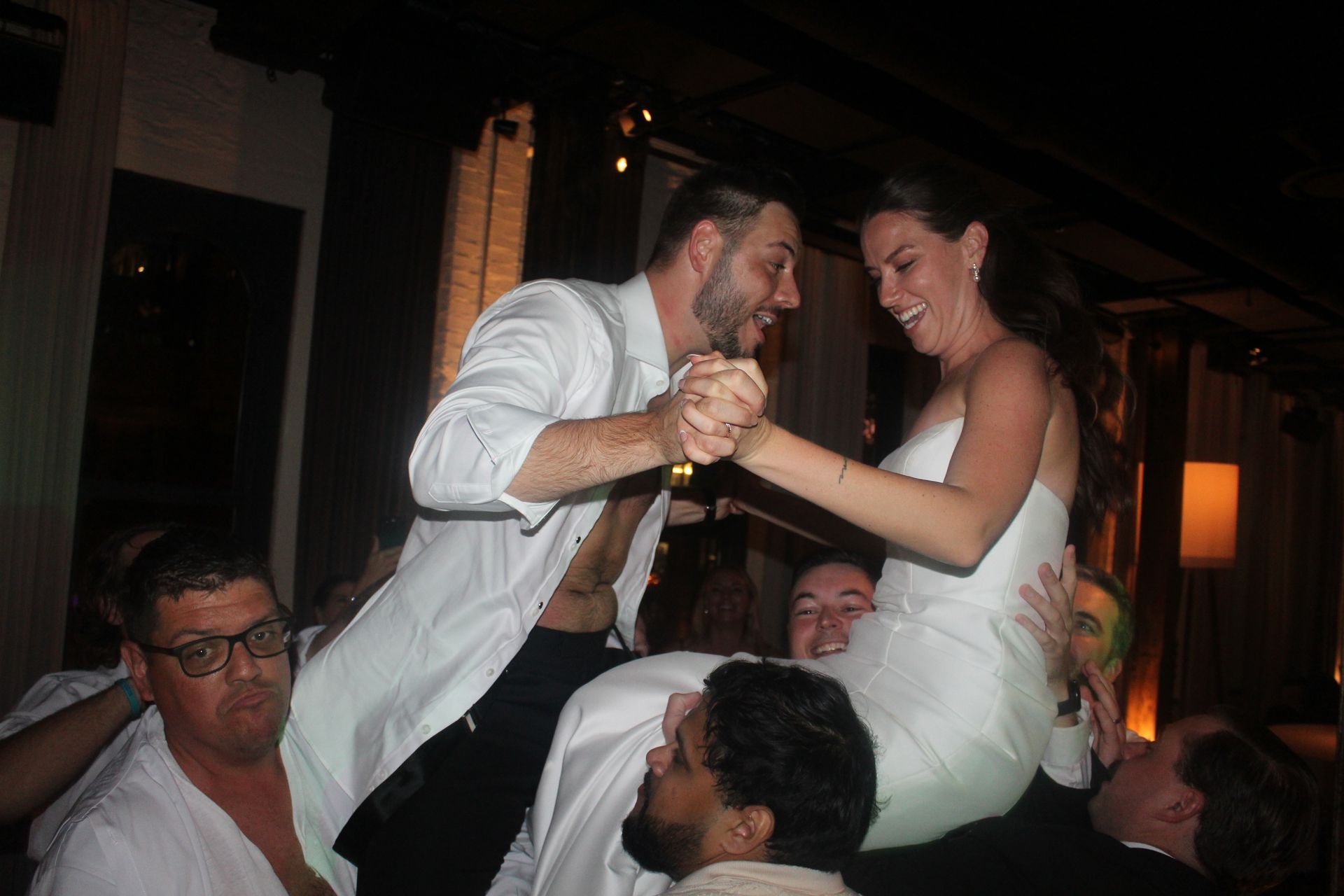 Bride and groom dancing on chairs at a wedding reception; people lifting them, smiling.