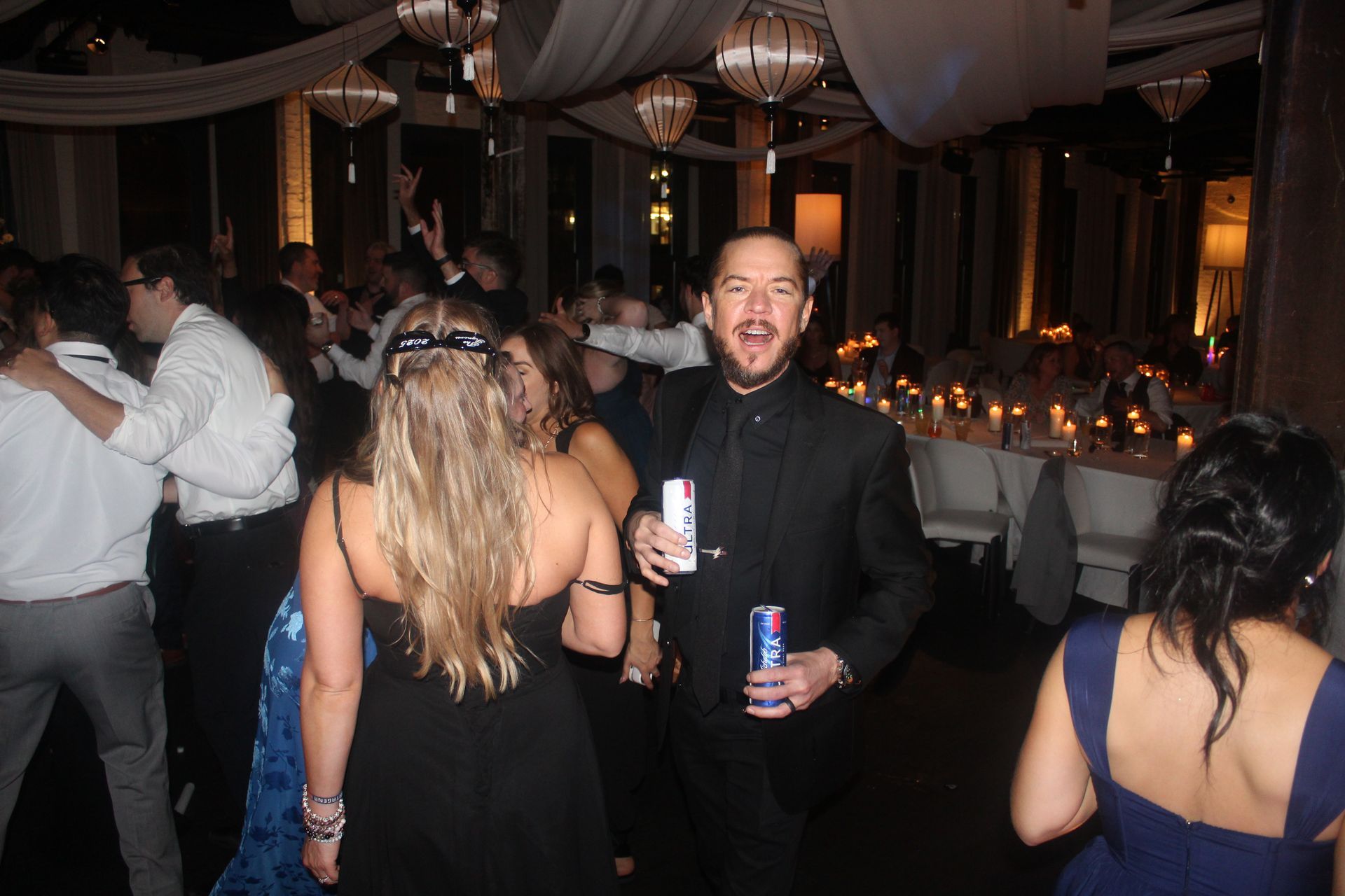 Party scene with dancing people, one man smiling with Red Bull cans. Dim, festive lighting.