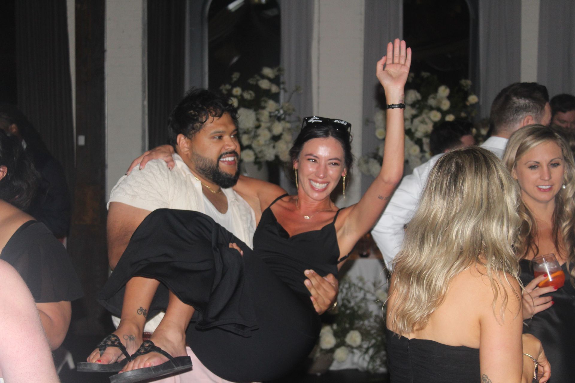 Man carrying a woman at a party; woman has arm raised. People in formal attire in a room with white flowers.