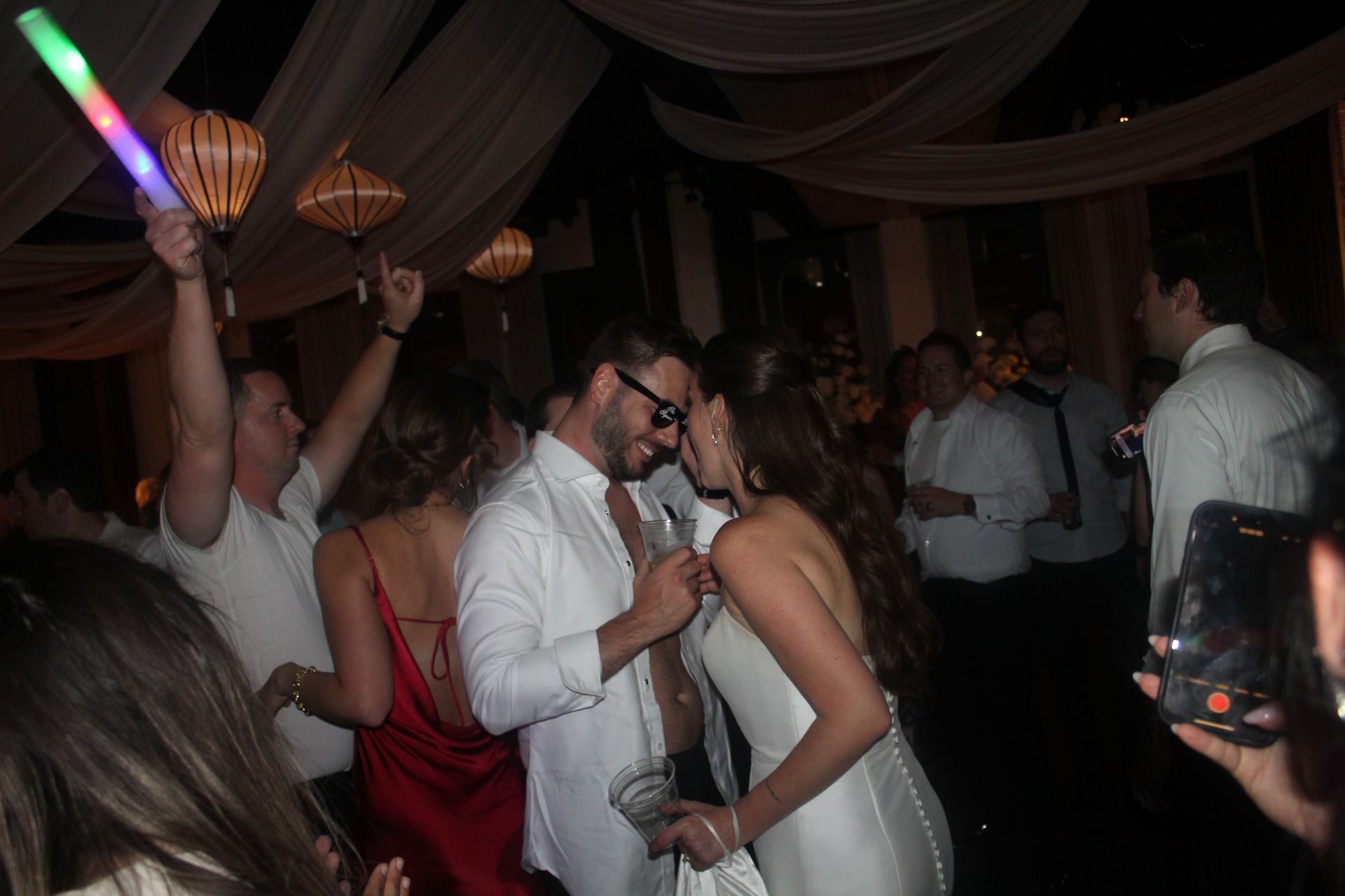 Couple kissing at a party with people dancing and holding lights; white and red attire.