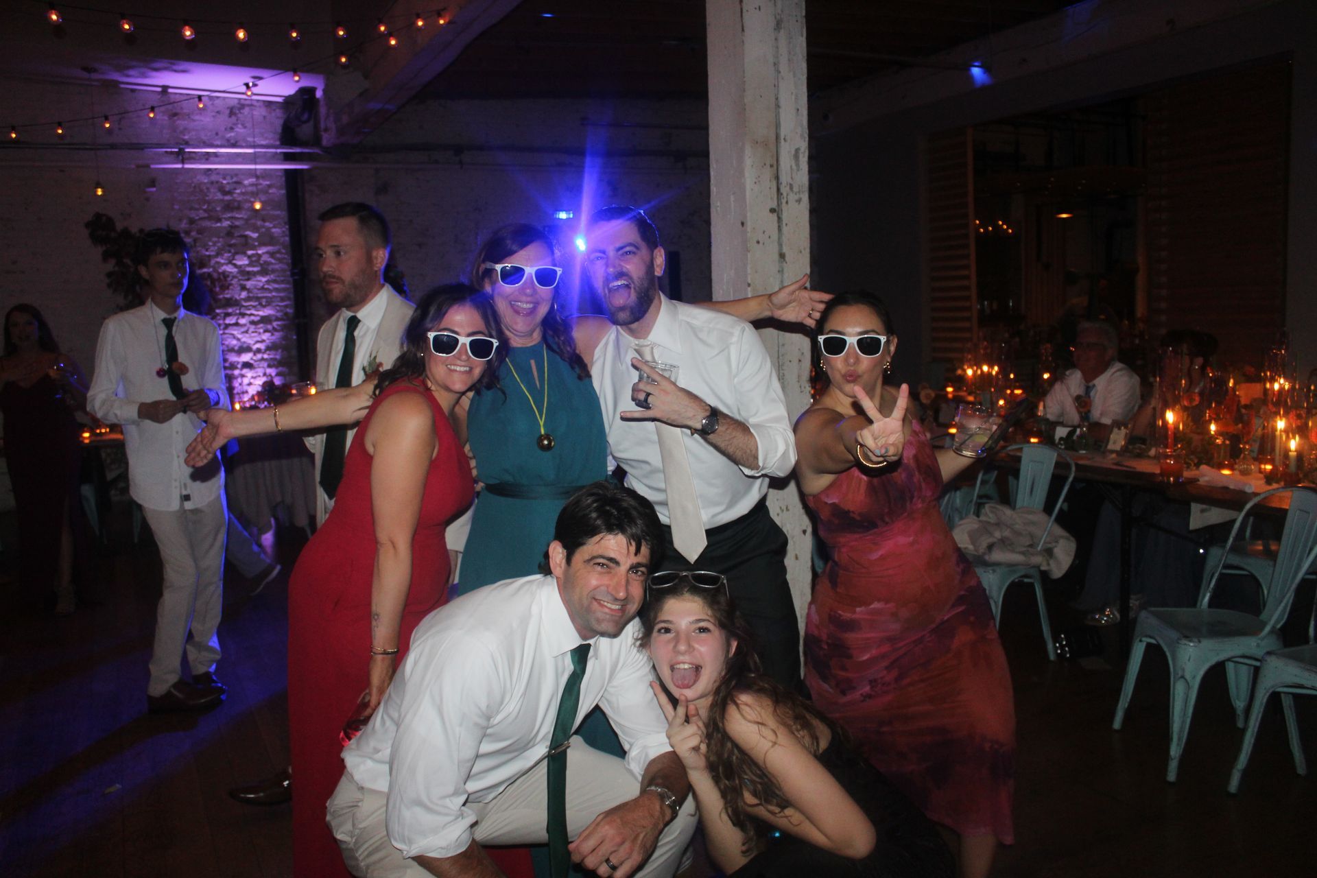 Group of people at a party, wearing sunglasses and smiling. Purple and blue lighting, indoor setting with tables.
