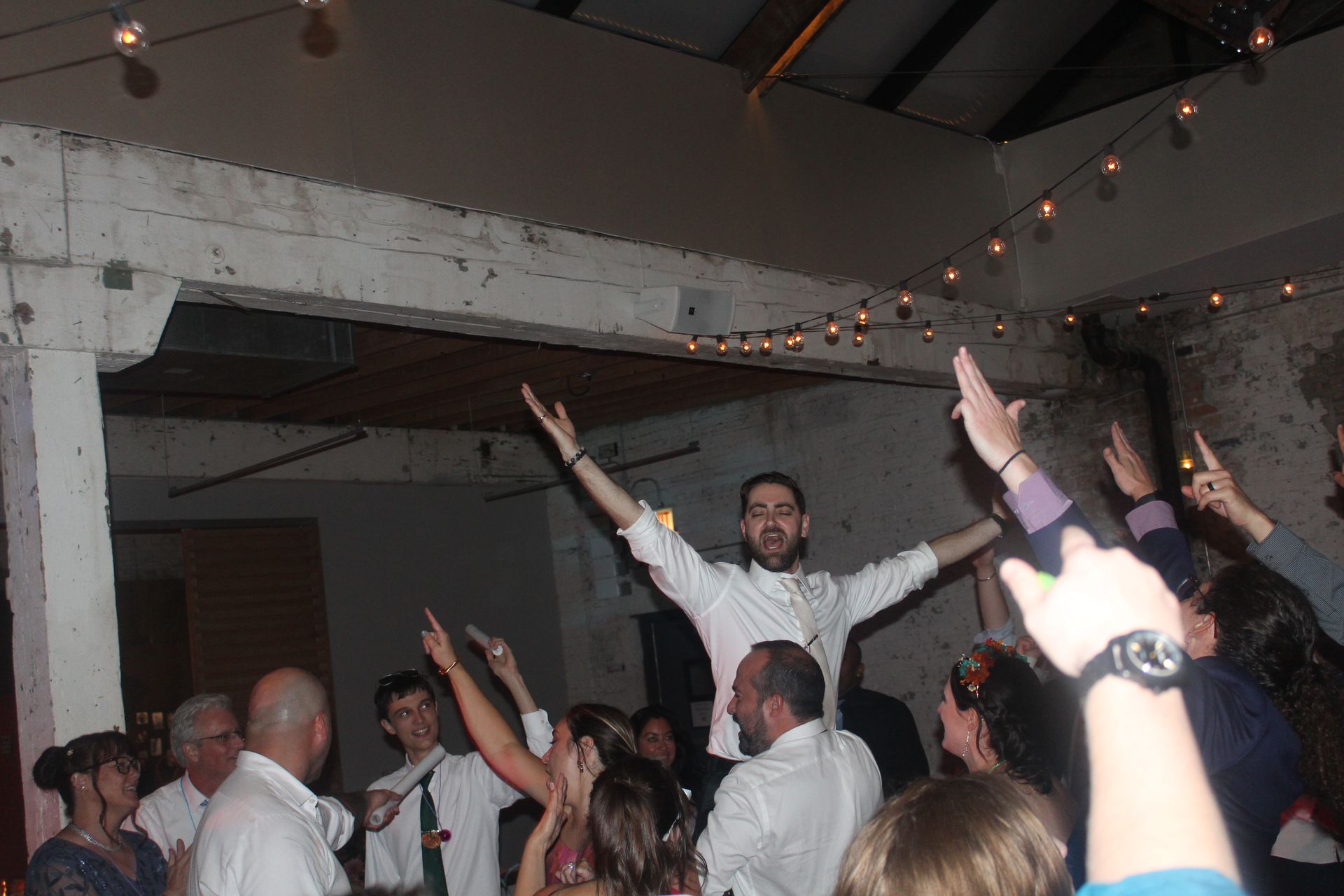Man standing with arms raised, surrounded by people with arms raised, lit by string lights in a room.