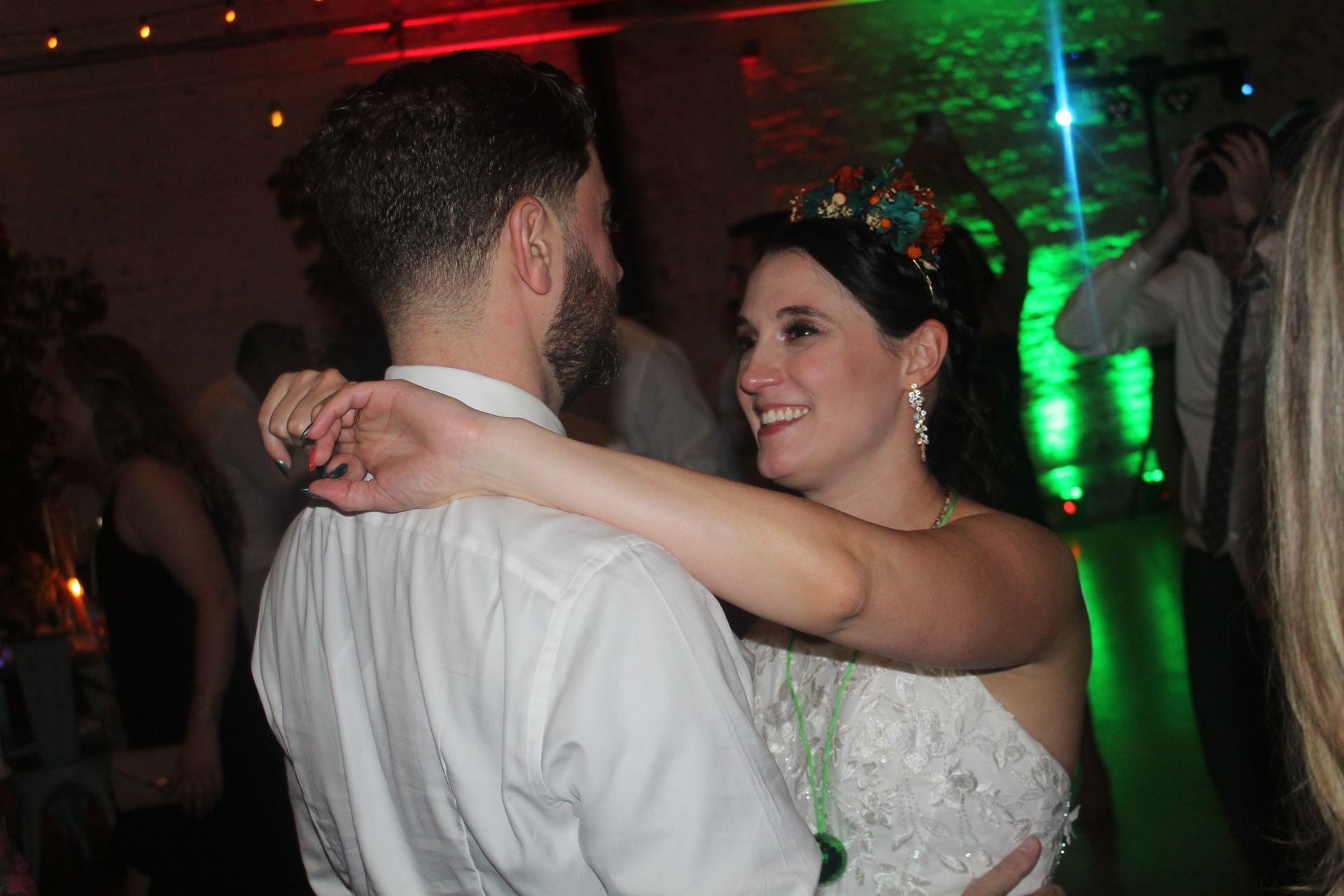 Couple dancing at a wedding reception; bride wearing crown, smiling at groom. Colorful lights in background.