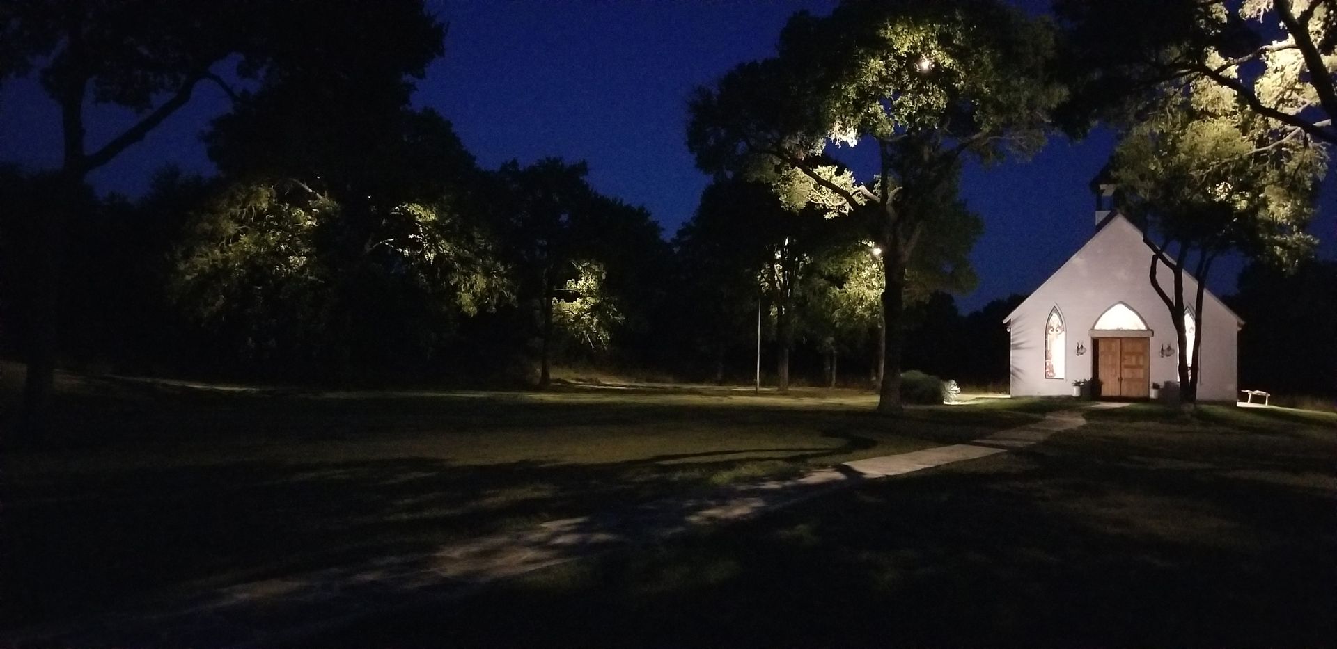 Nighttime view of a small white chapel lit by warm lights, surrounded by trees and a dark park