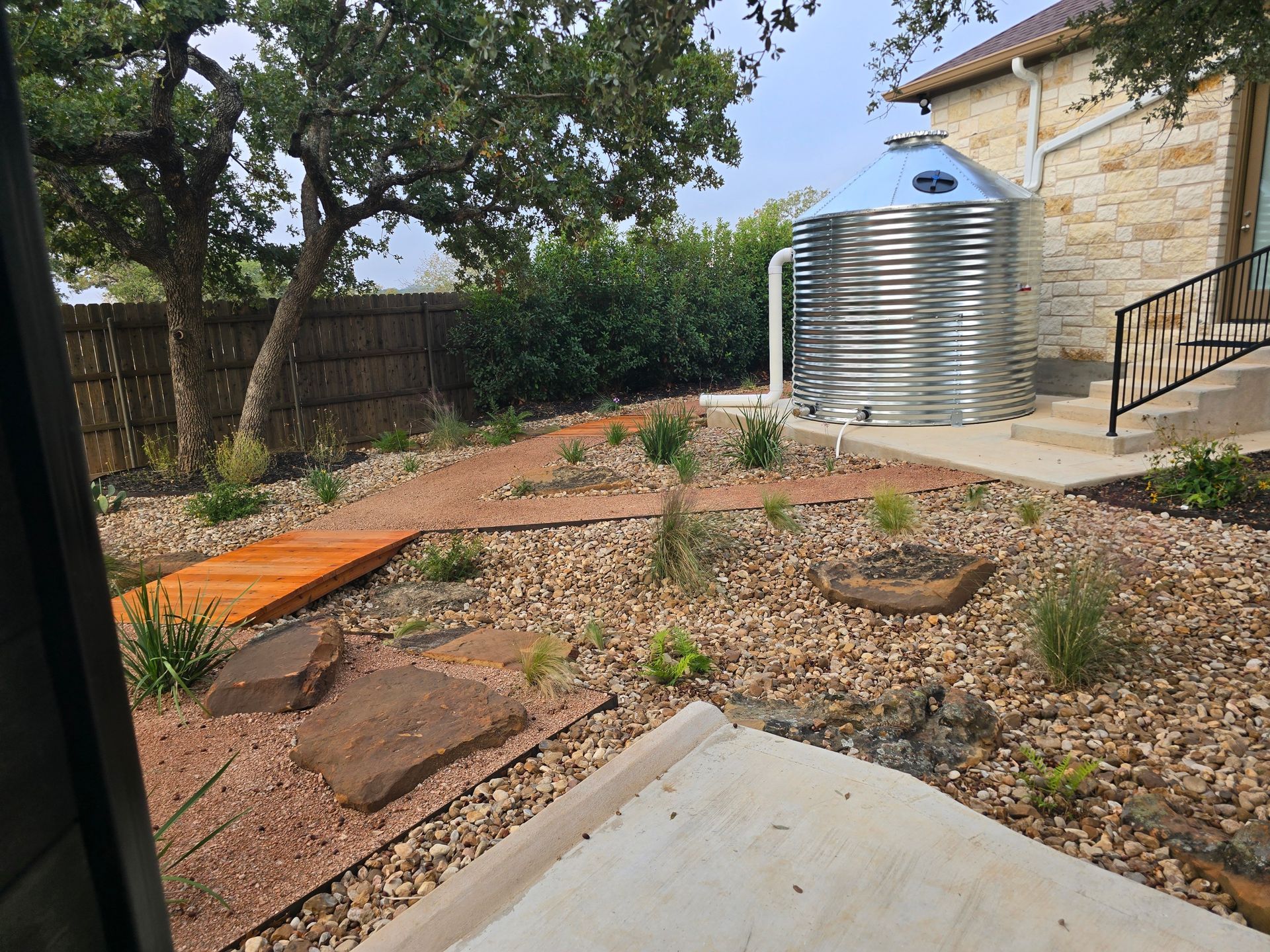 Rocky backyard garden with shrubs, trees, a wooden fence, and a large metal water tank beside a stone house.