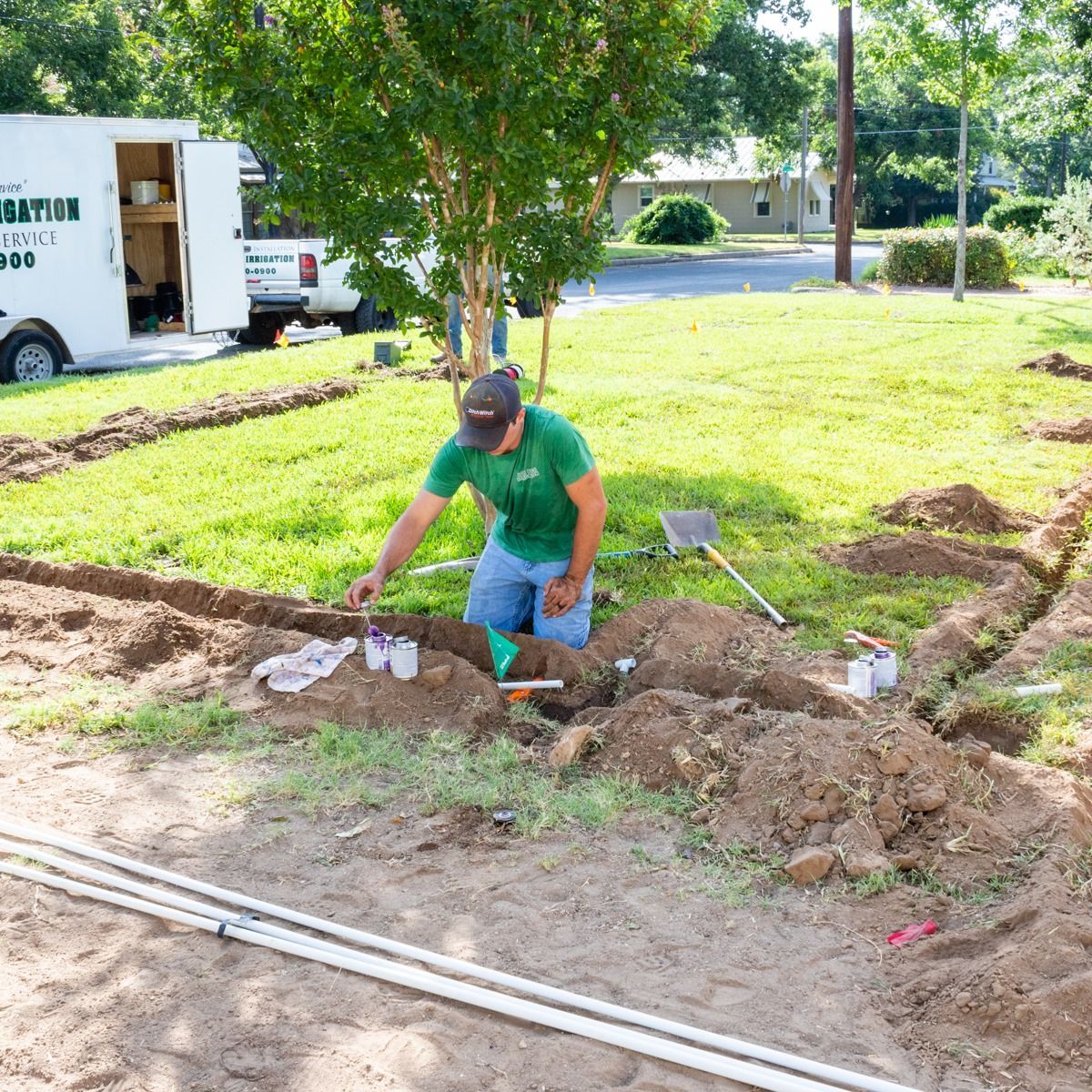 Worker shoveling dirt beside a trench in a grassy yard, with construction materials nearby.