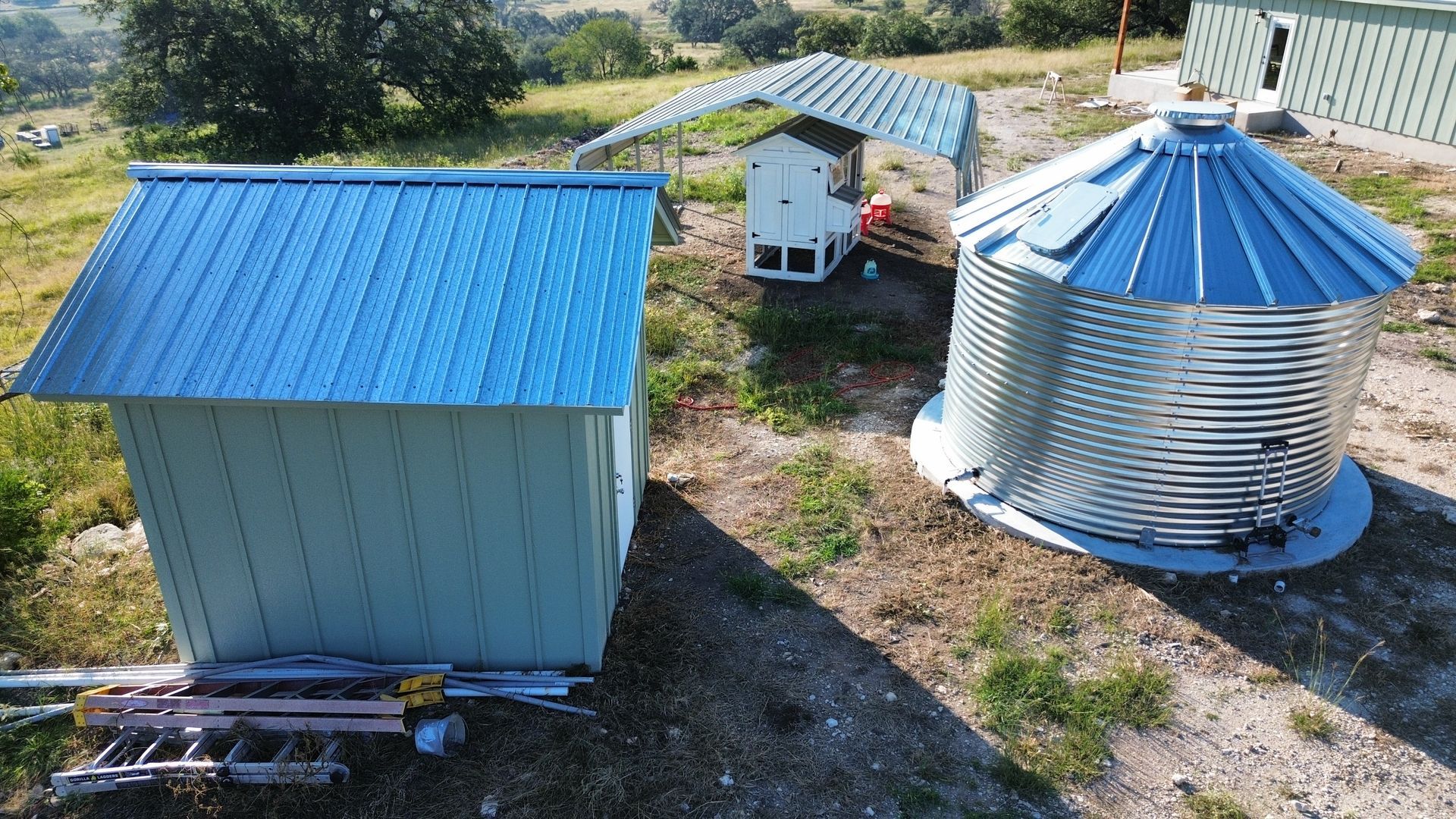 Aerial view of small rural buildings and a large blue-roofed metal grain silo on dirt ground