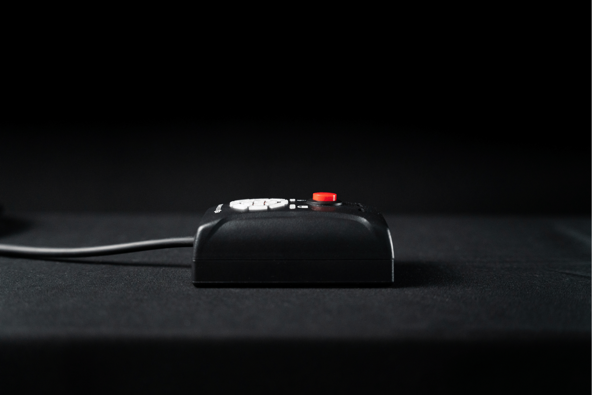 Black rectangular device with red button on top, connected to a wire, sitting on black surface.