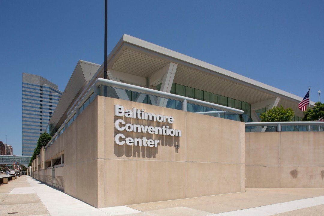 Exterior view of the Baltimore Convention Center, a tan stone building with large glass windows and a US flag on a pole.