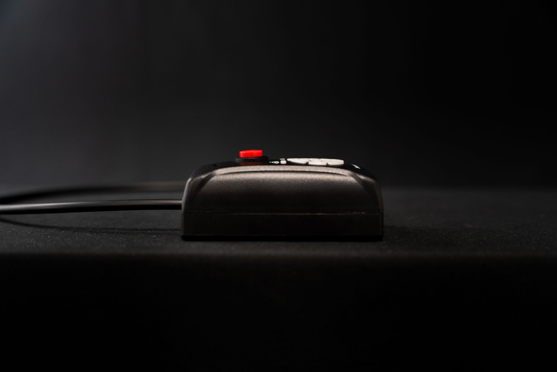 Black rectangular device with red button on top, connected to a wire, sitting on black surface.