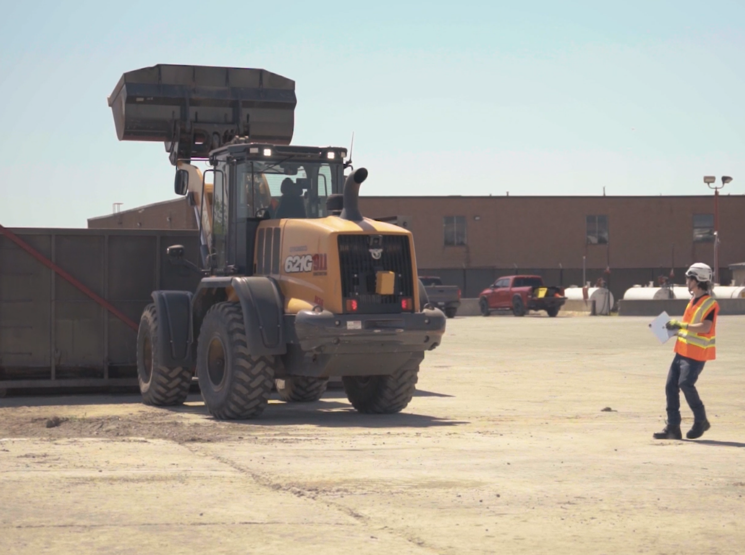 A person in a high-visibility vest stands near a yellow construction loader while holding a clipboard outdoors.