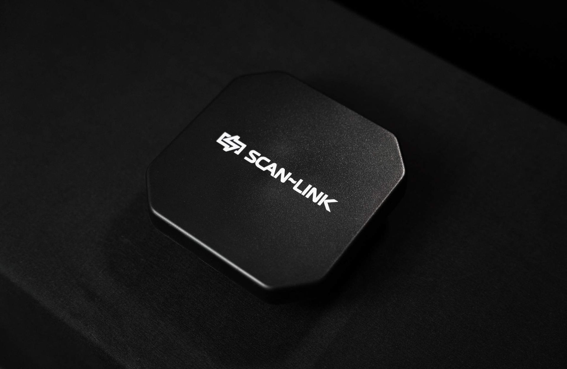 Black, octagonal Scan-Link device on black fabric. White logo and text on top.