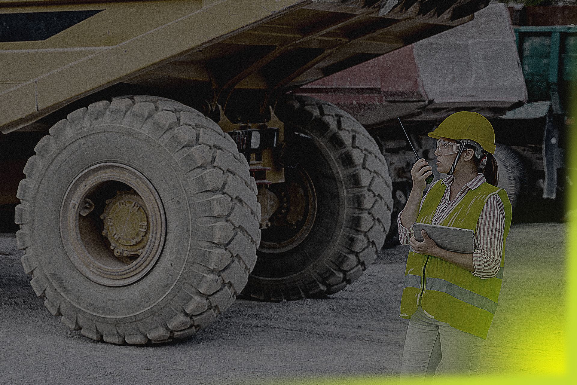 A woman is standing in front of a large dump truck talking on a walkie talkie.