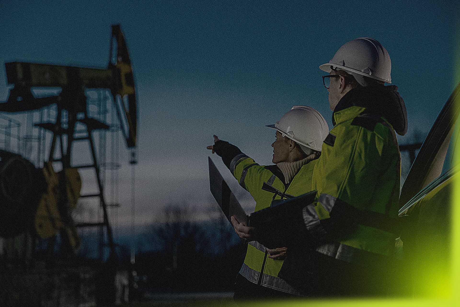 Two oil workers are standing in front of an oil pump at night.