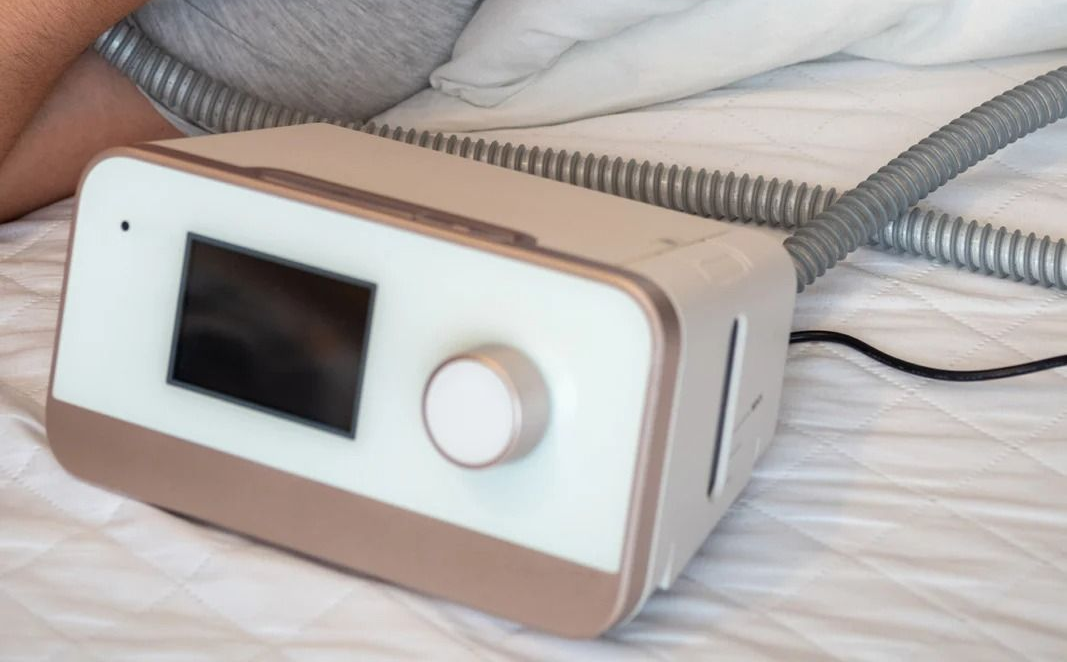 A bedside CPAP machine with a digital display and a control dial, connected to a flexible air hose on a white sheet.