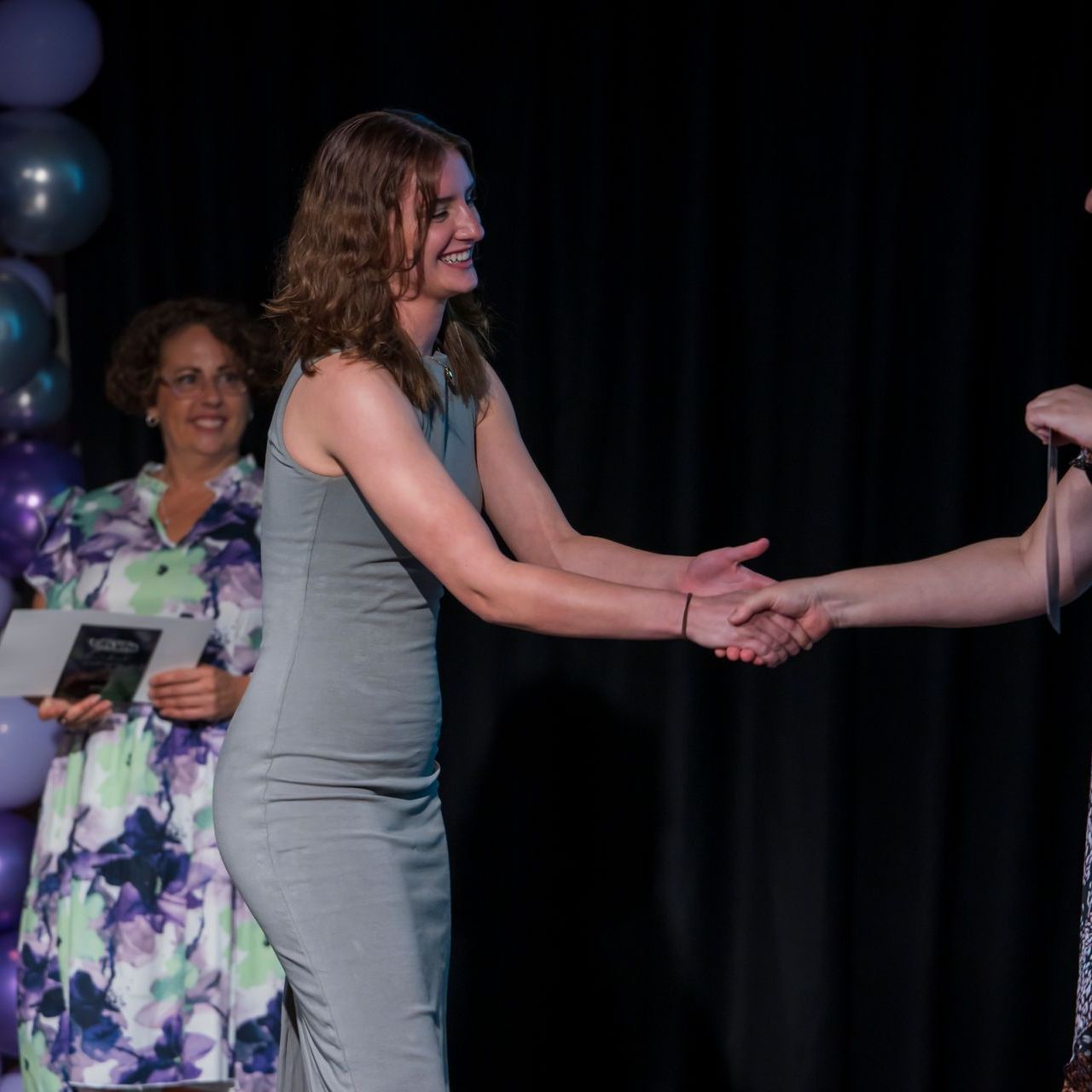 Two women are shaking hands on a stage with balloons in the background.
