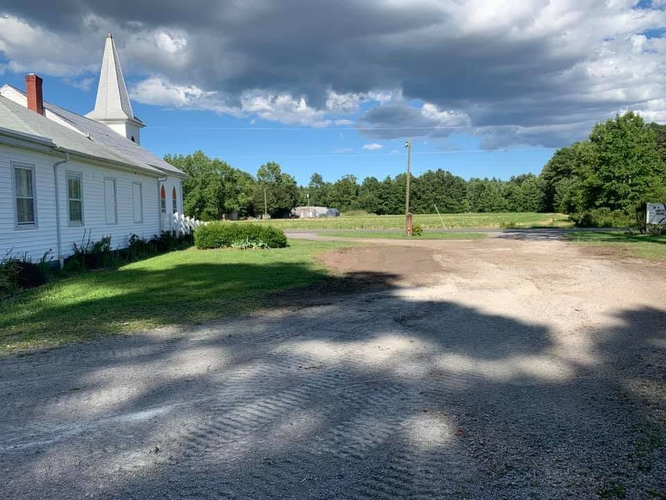 White church beside a gravel lot under dramatic clouds, with green trees and grass in the background