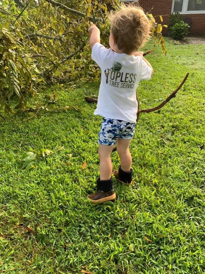 Child in a white shirt and blue shorts reaching up in a grassy yard near bushes.
