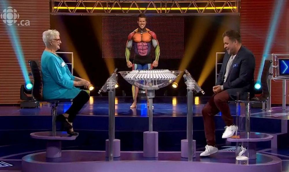 Un homme en costume de super-héros se tient devant une table avec deux personnes assises autour.