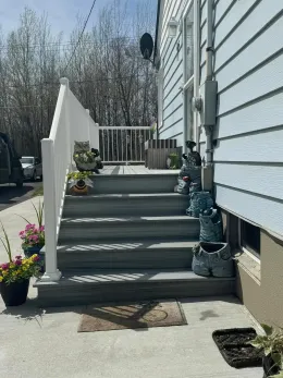 A set of gray wooden porch steps leading to a light blue house, decorated with several potted plants and garden figures.