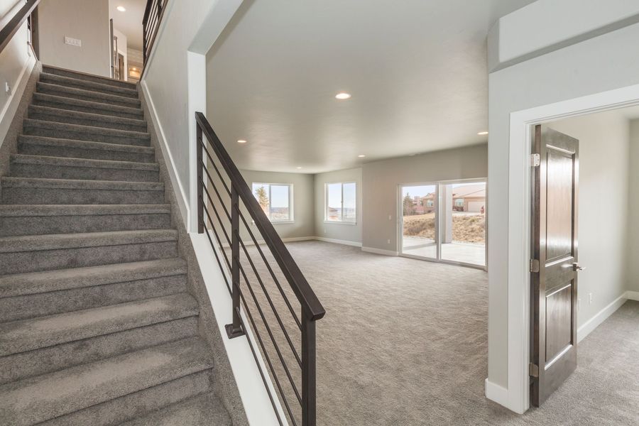 A gray-carpeted staircase with dark railings next to an open, carpeted living area with light-colored walls.