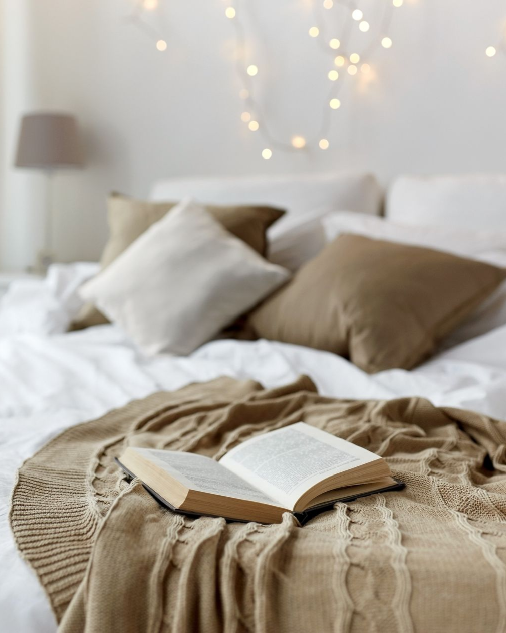 An open book on a tan blanket, layered on a bed with pillows, fairy lights in background.