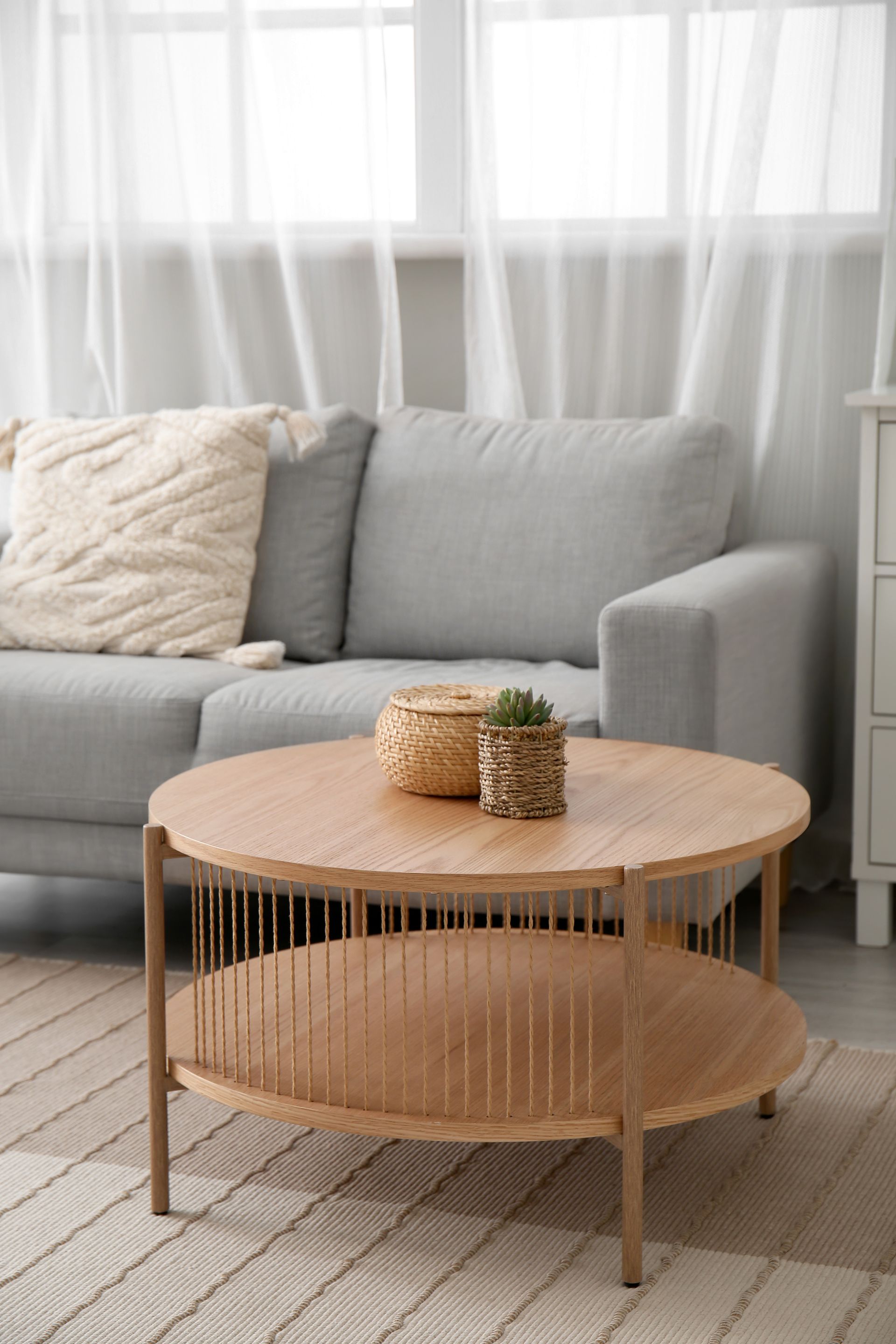 Round wooden coffee table with woven basket and plant in front of a gray couch.
