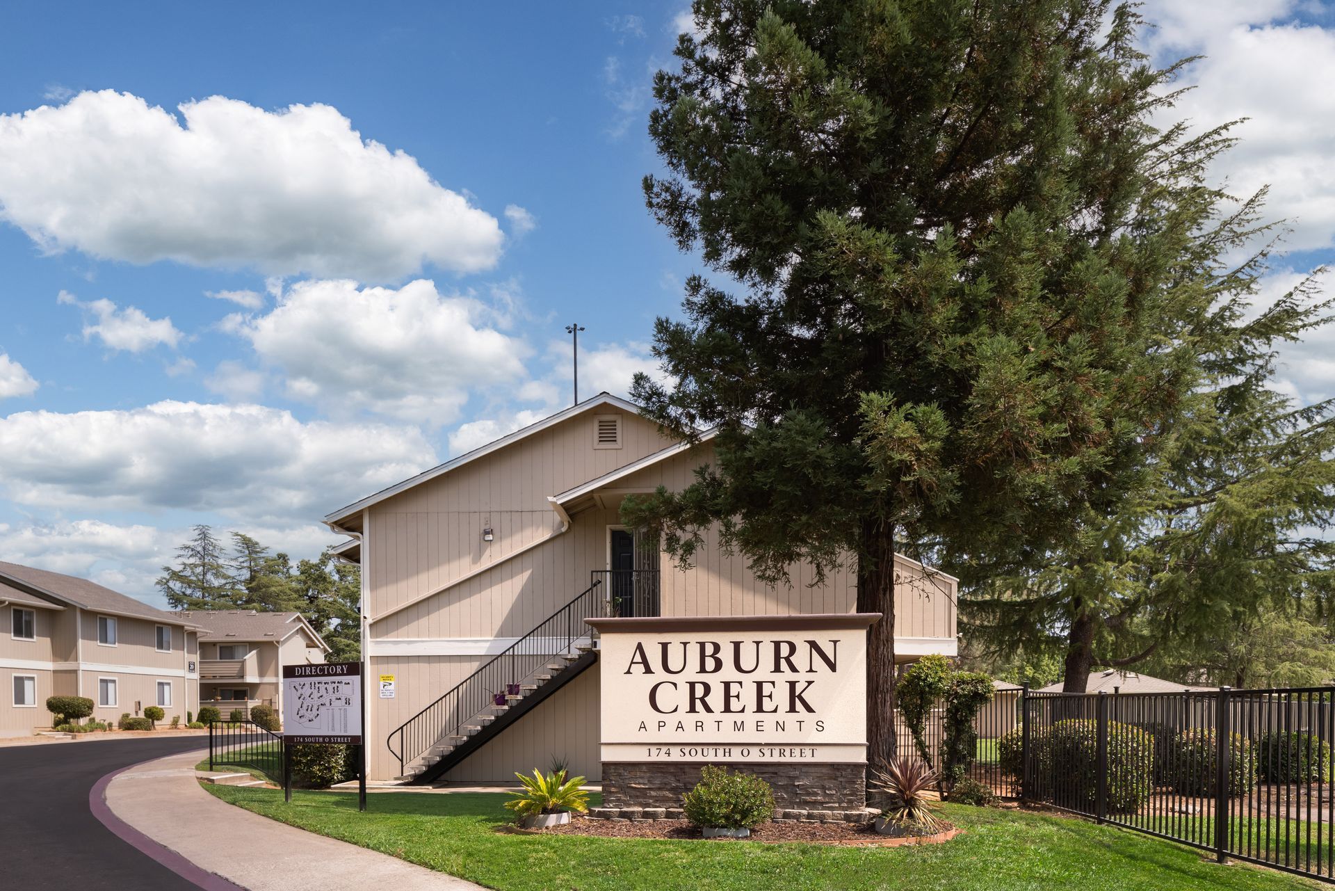 Auburn Creek apartment complex sign under a tree on a sunny day; buildings in the background.