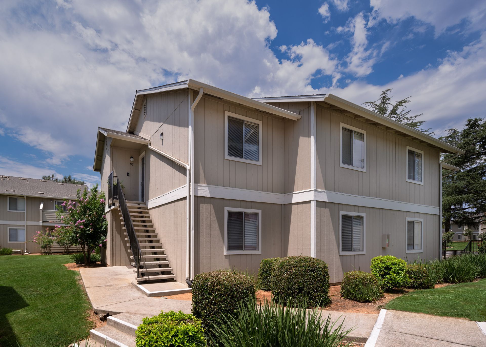 Two-story beige apartment building with outdoor stairs, bushes, and a green lawn.