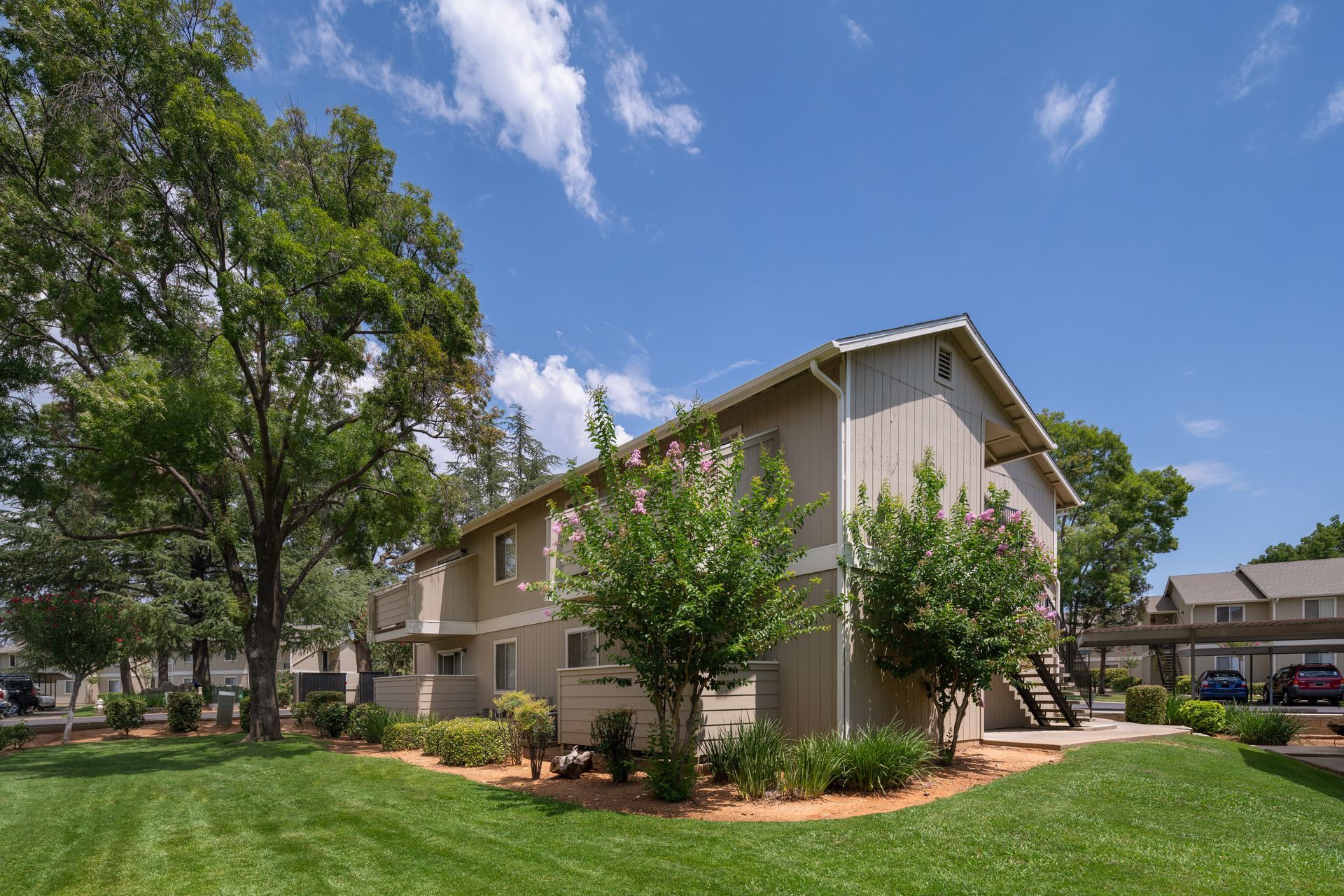 Two-story tan apartment building with green lawn and trees under blue sky.